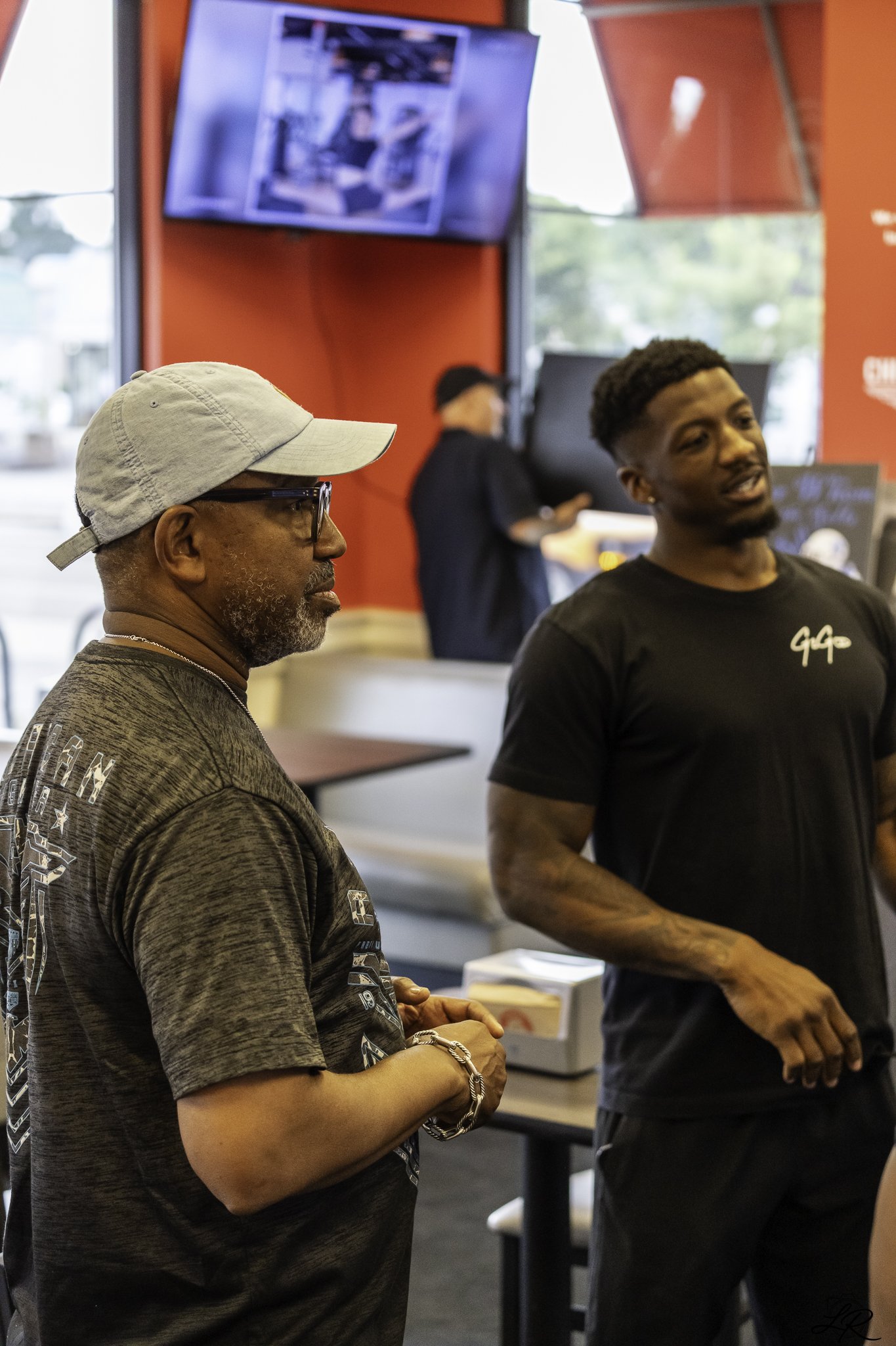 Two men standing inside a fast-food restaurant near a counter, with a red wall and a TV screen behind them. One man is wearing a beige cap and glasses, the other has a short fade hairstyle and a black T-shirt.