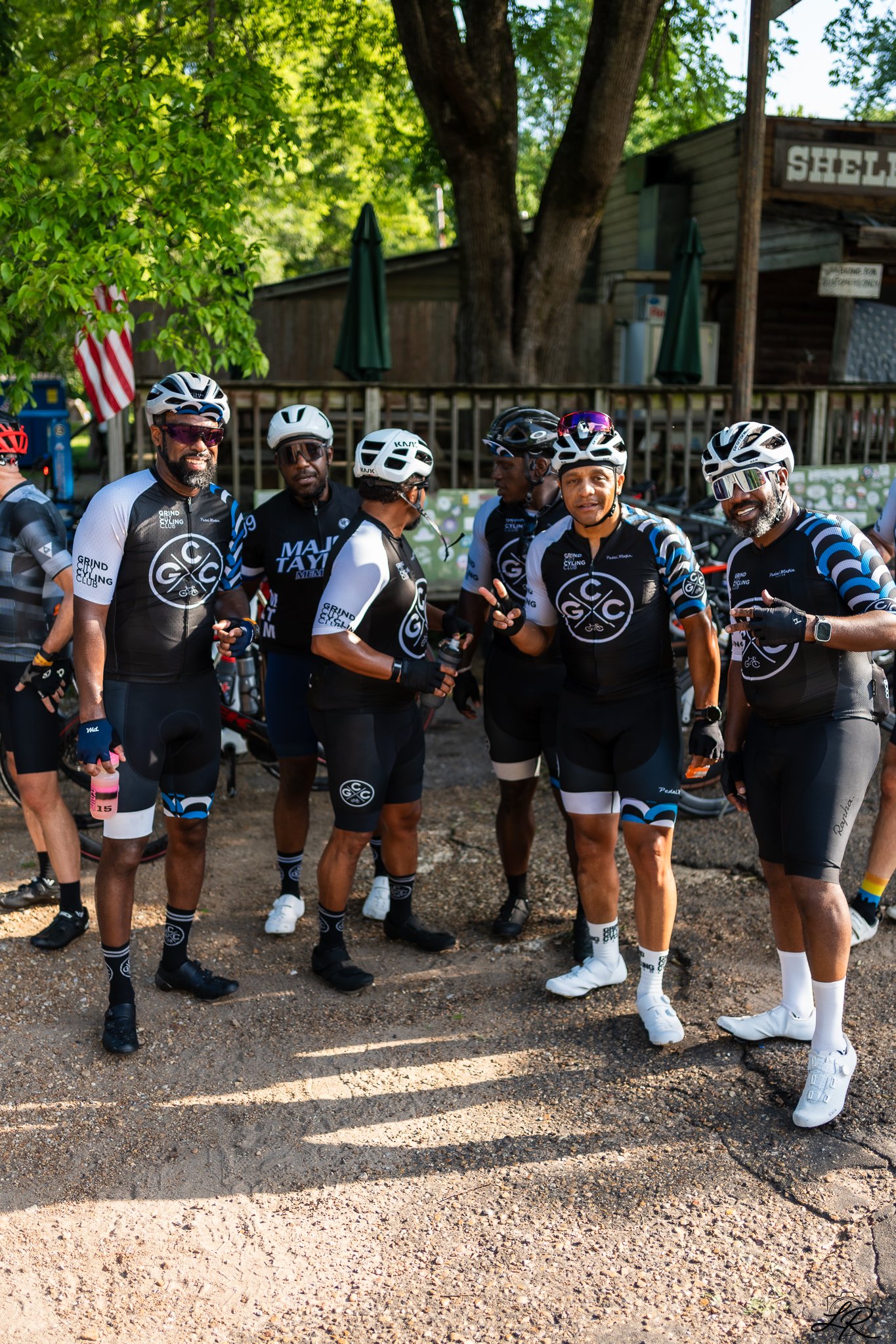 A group of six men wearing matching black and blue cycling jerseys and helmets, standing outdoors in a wooded area, posing for a photo with some smiling and others looking at each other.