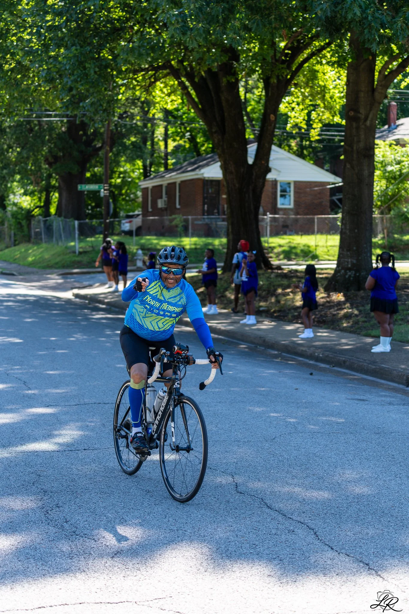 A man wearing a blue sports outfit and helmet is riding a black road bike on an urban street. In the background, there are children in blue uniforms cheering and standing near a park with large leafy trees and a brick house.