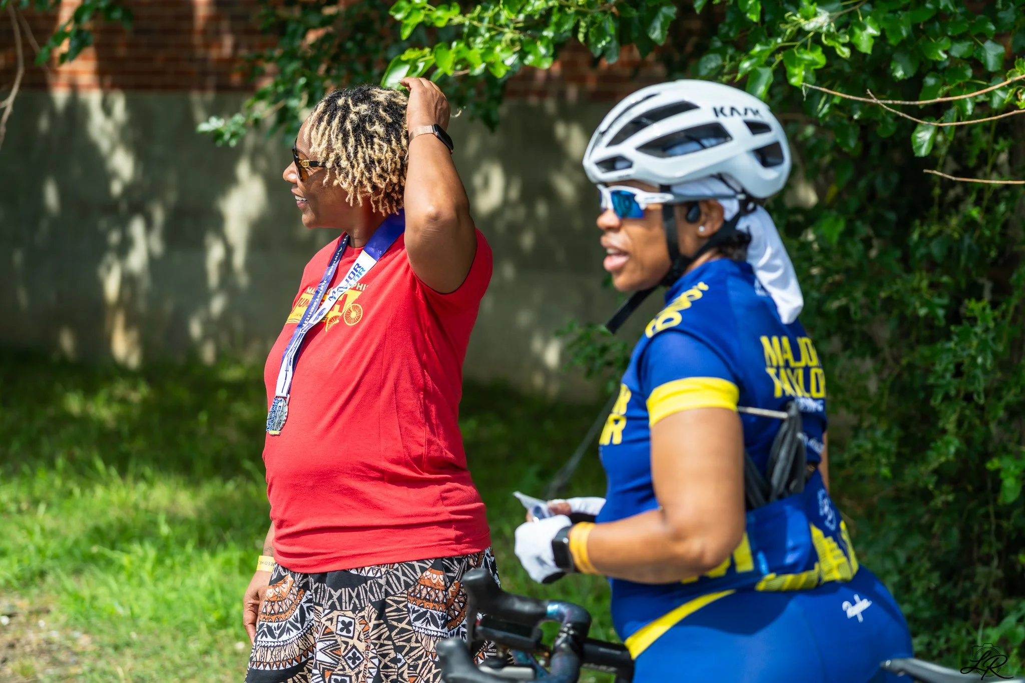Two women outdoors, one in a red shirt with a medal, and the other in a blue cycling outfit with a white bike helmet, smiling and talking with greenery and a brick wall in the background.