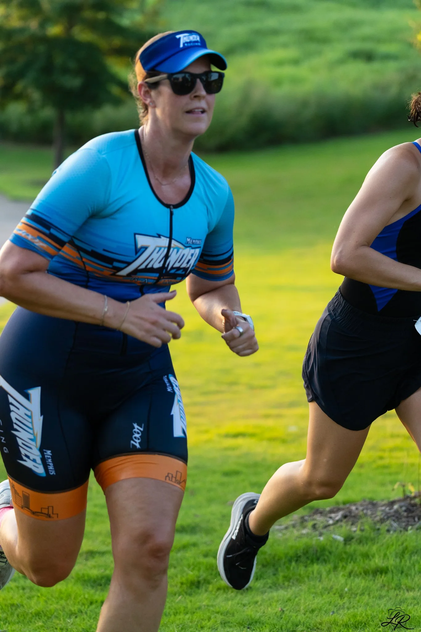 Two women running outdoors on a grassy field, wearing athletic clothing and sunglasses, with a blurred green background.