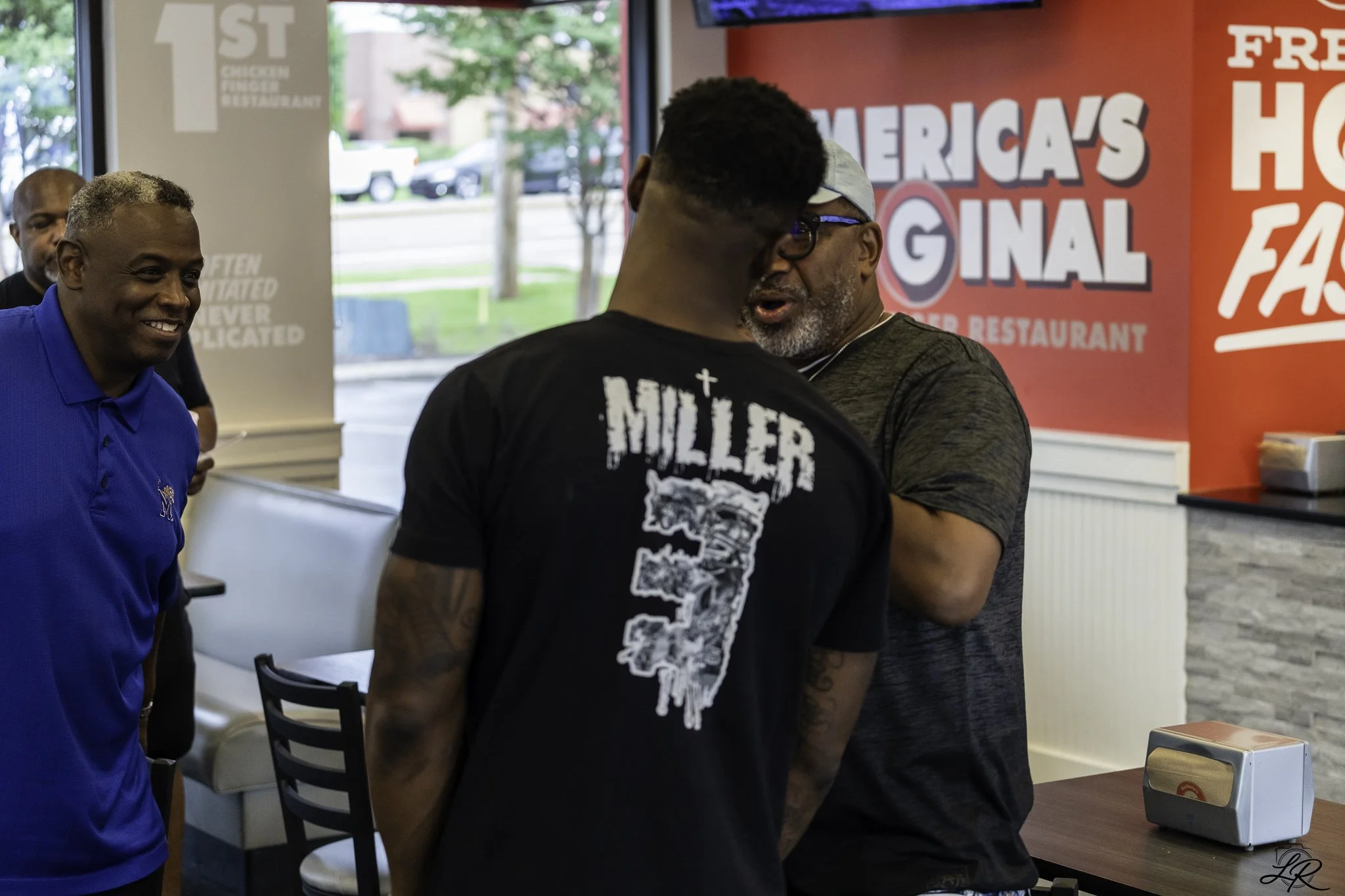 Three men are in a restaurant near the counter, with one man in a blue shirt smiling on the left, and two men in black and gray shirts facing each other in the center. They are engaged in a conversation, with the man in gray wearing glasses and a cap