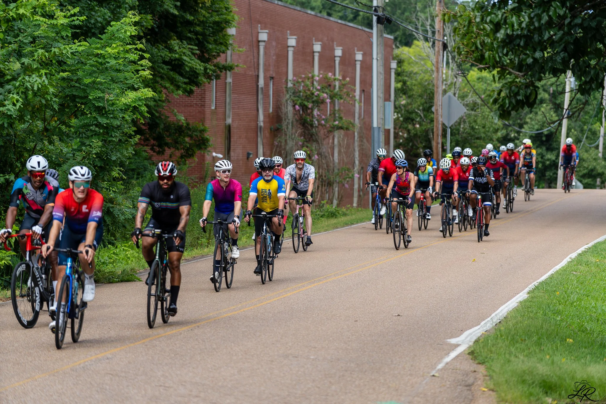 A large group of cyclists riding on a paved road surrounded by green trees and bushes. There is a brick building in the background with some pink flowering plants climbing on it. The cyclists are wearing helmets and colorful sports attire.