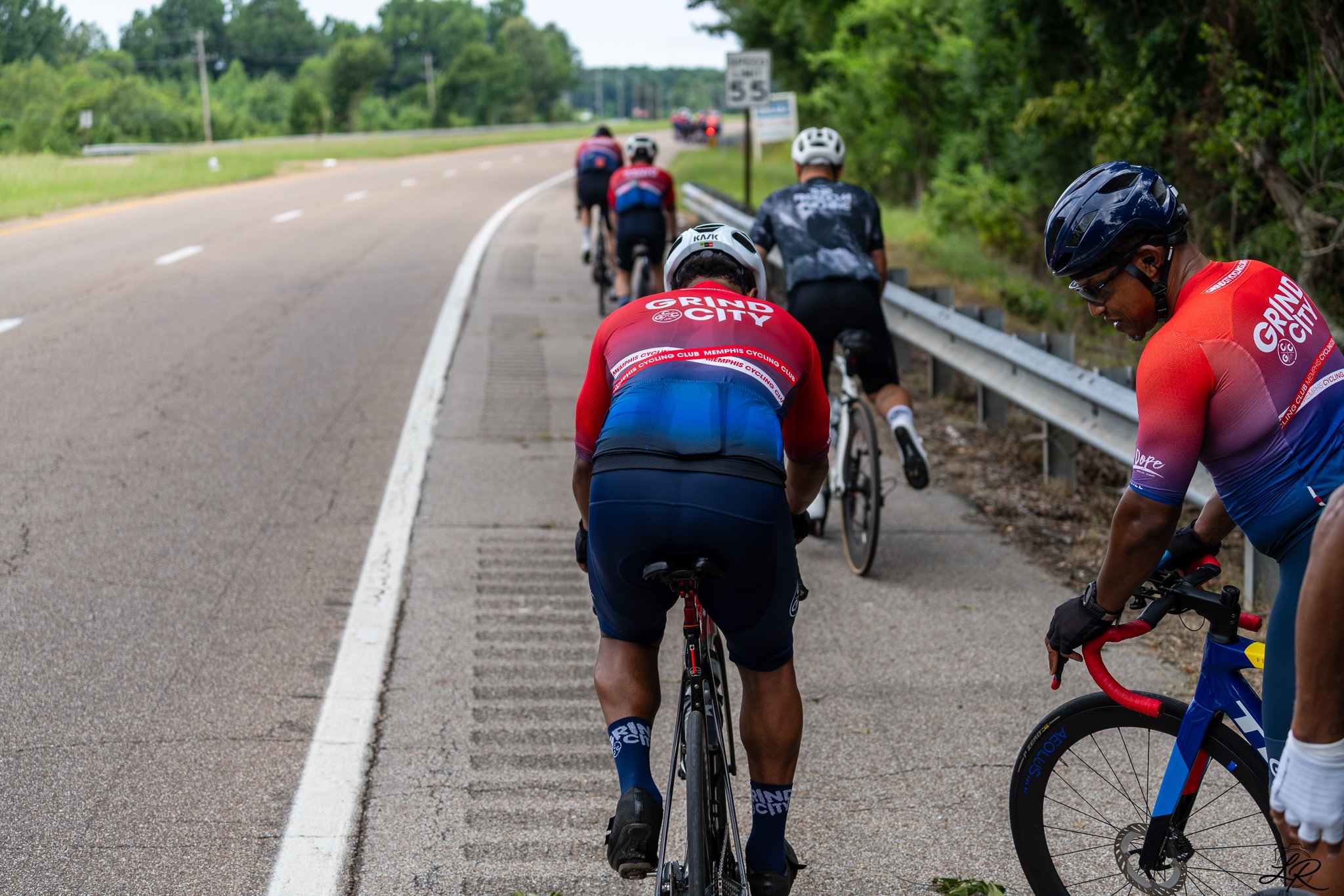 Group of cyclists riding on the side of a rural road, some wearing red jerseys and helmets, with green trees and a road sign in the background.