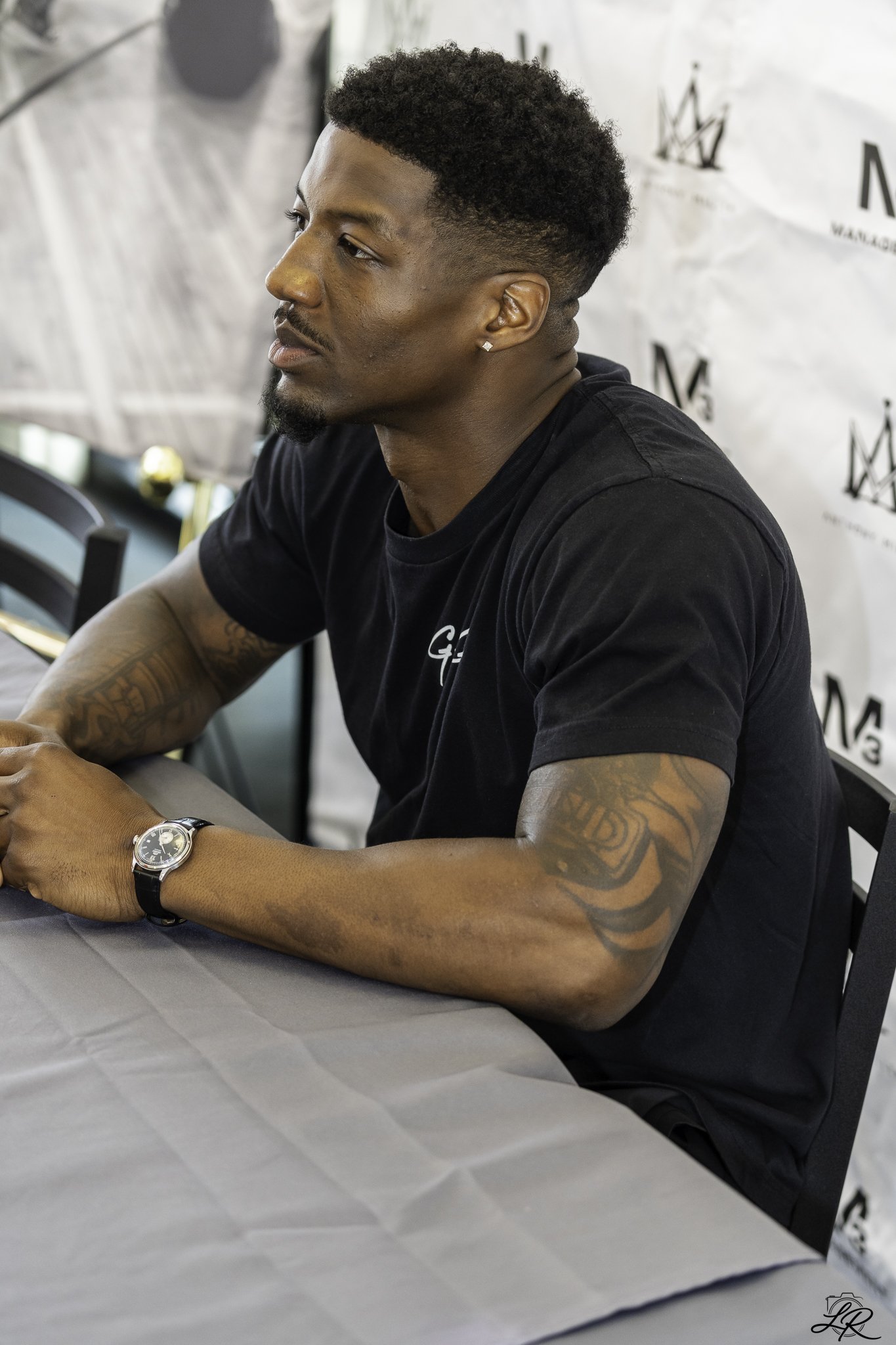 A young man with dark skin, curly hair, a beard, and earrings, wearing a black t-shirt, sitting at a table with a gray tablecloth. He has a silver wristwatch on his left wrist and tattoos on his right arm. The background includes a white wall with bl
