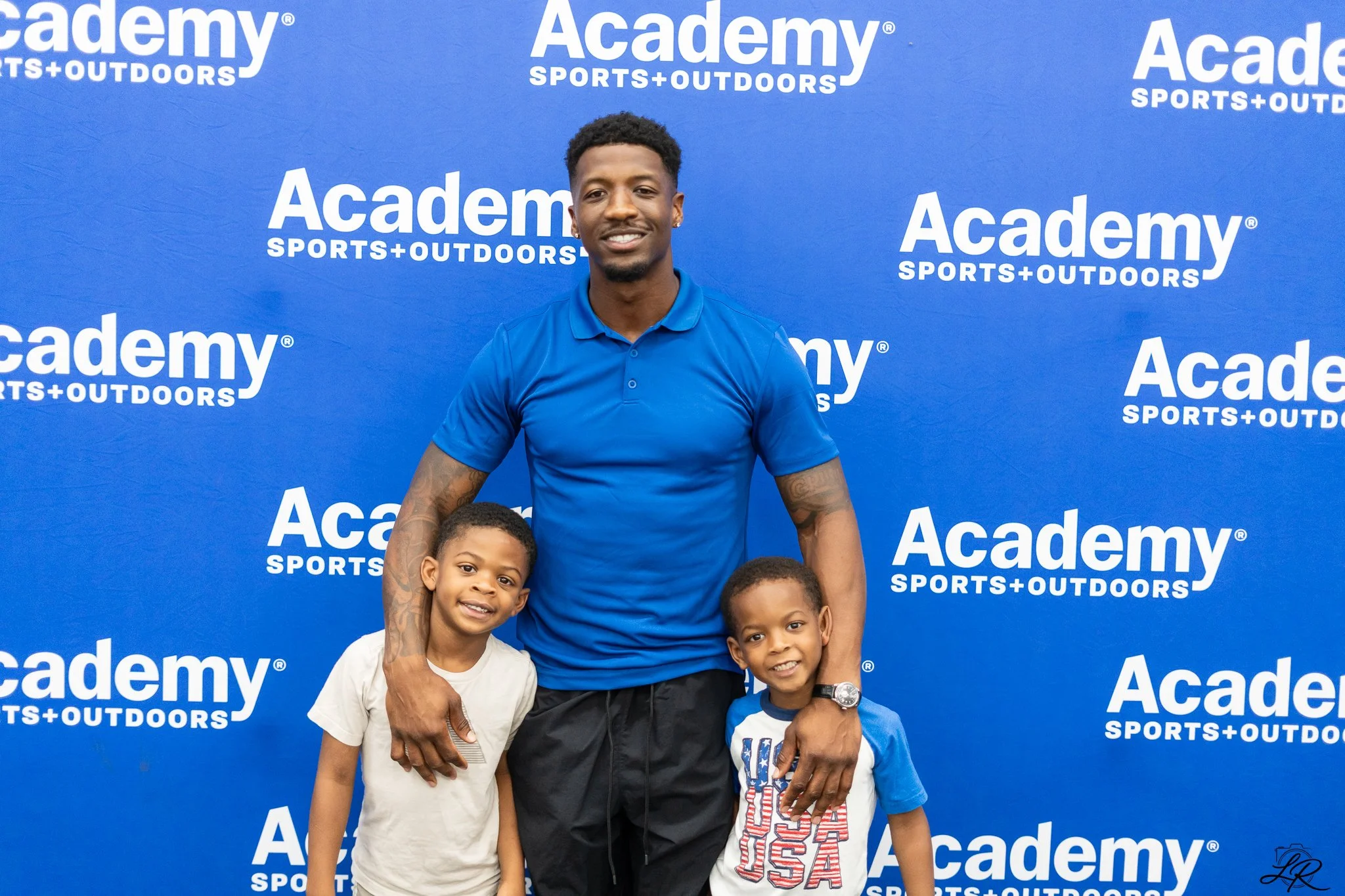 A man smiling and posing with three young boys in front of a blue backdrop with the text 'Academy Sports + Outdoors' repeated multiple times. The man is wearing a blue shirt, and the boys are dressed casually, one in a white shirt, another in a blue 