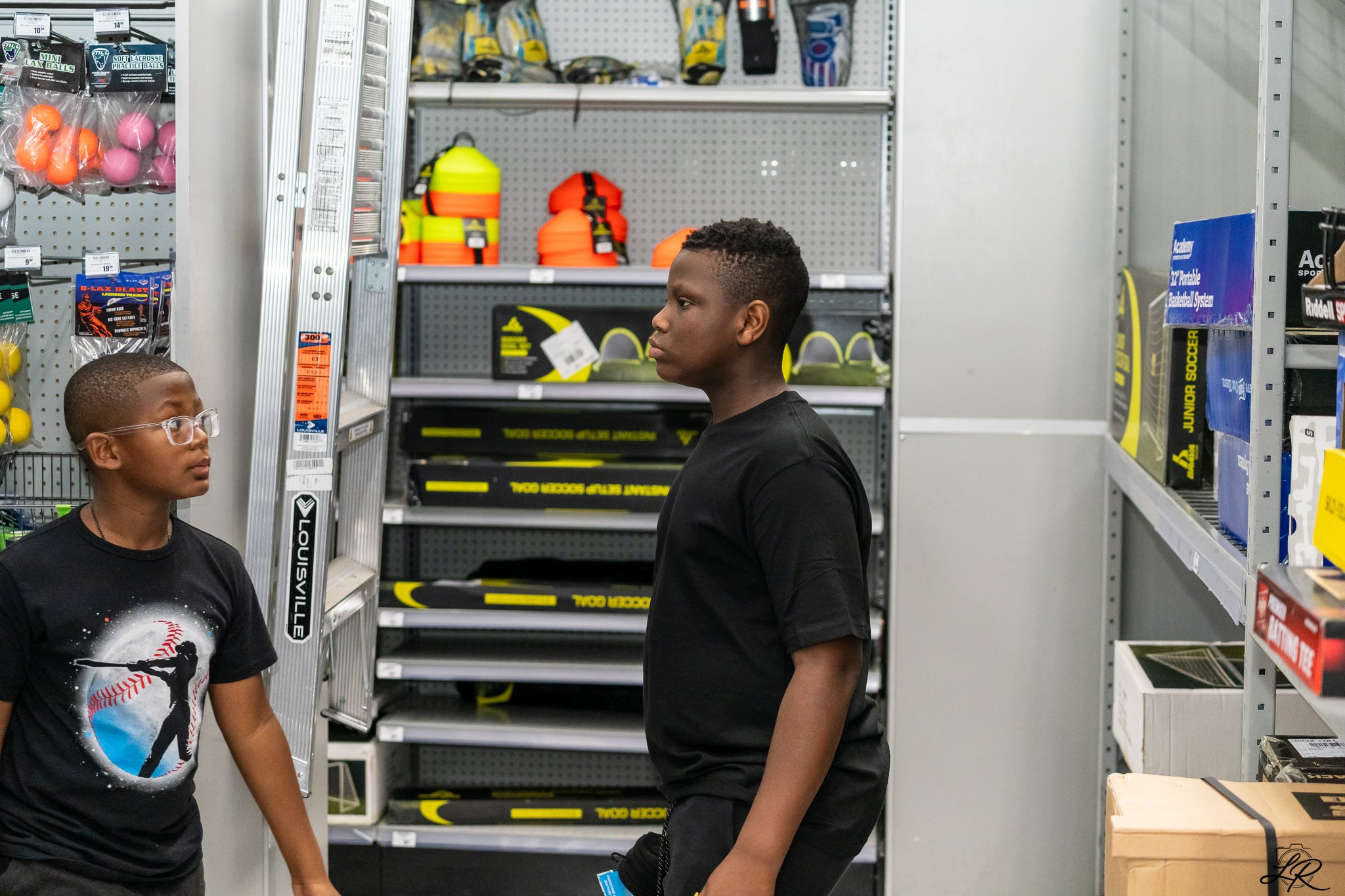 Two young boys standing in a sporting goods store, surrounded by shelves with sports equipment such as balls, bags, and hockey sticks. One boy with glasses and a black t-shirt is looking at the other boy, who has short curly hair and a black t-shirt,