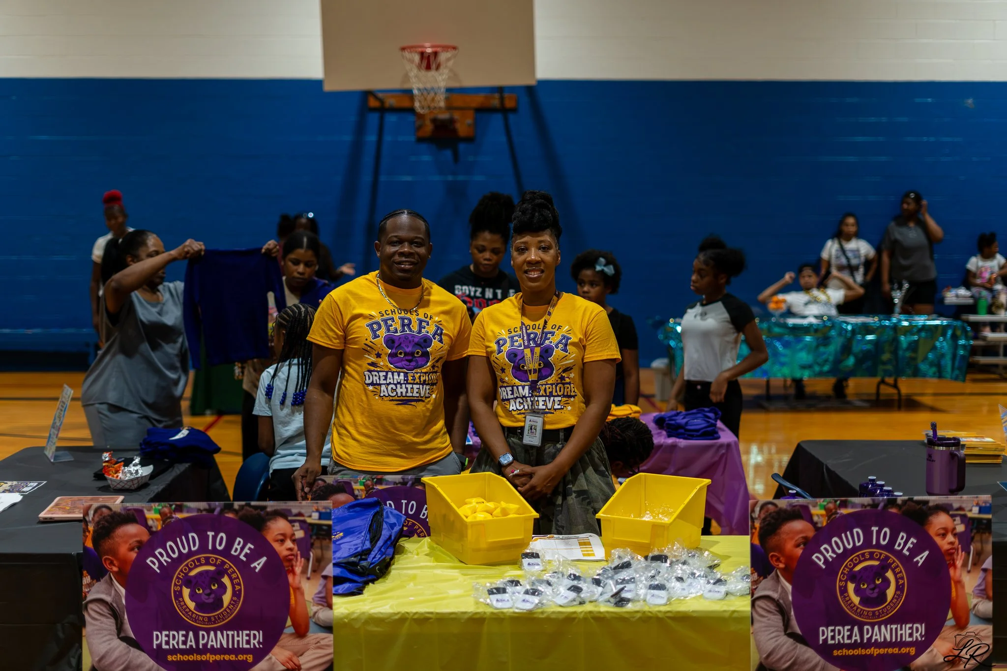 A group of children and adults inside a gymnasium participating in a school event. Two individuals in yellow shirts stand in front of a table with purple and yellow items, with the shirts branded "Proud to be a Perea Panther." Children hold large pur