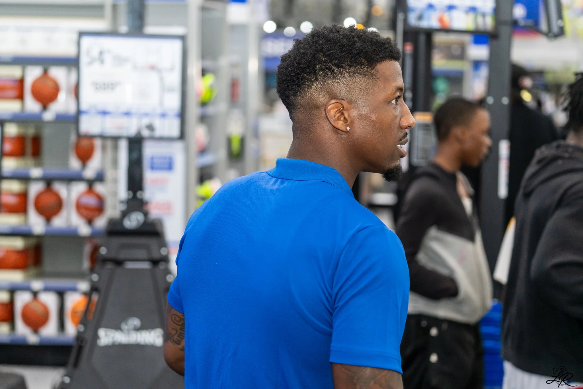 Side profile of a young man in a blue shirt with short curly hair and earrings standing inside a store, with people and store shelves in the background.