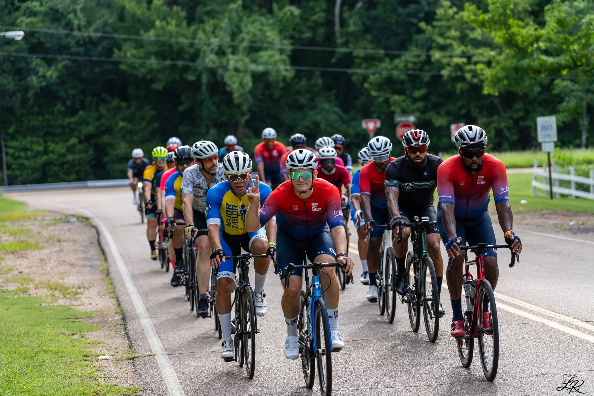 A large group of cyclists riding on a paved road through a lush, green, wooded area, with some making peace signs and wearing colorful cycling jerseys and helmets.