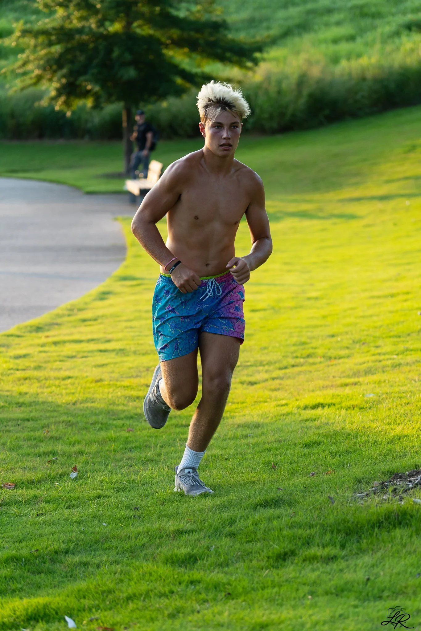 A young man with bleached blonde hair running outdoors on a grassy park path, shirtless, wearing colorful shorts and running shoes.