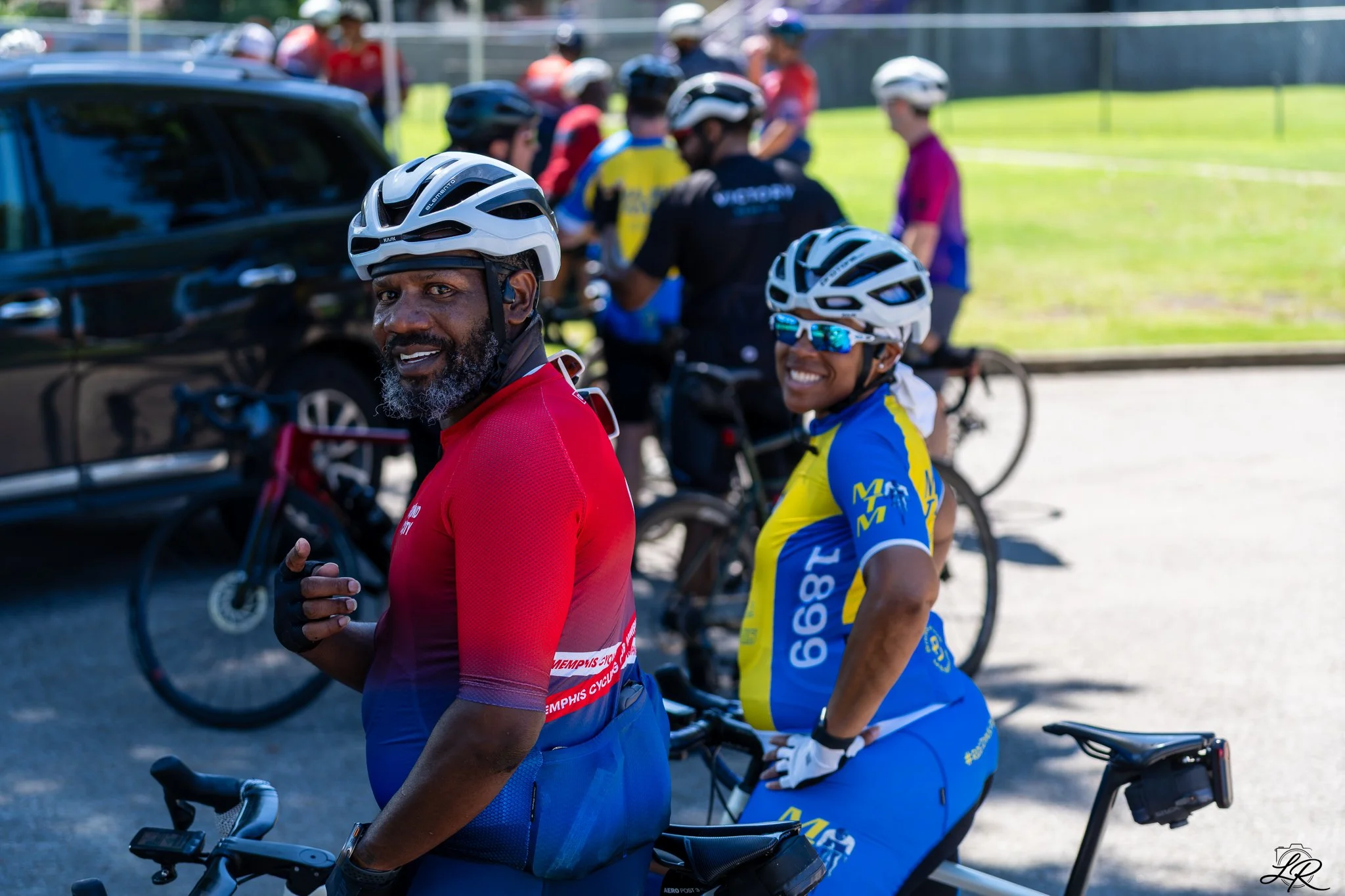 Cyclists wearing helmets and colorful jerseys gather outdoors near parked cars on a sunny day.