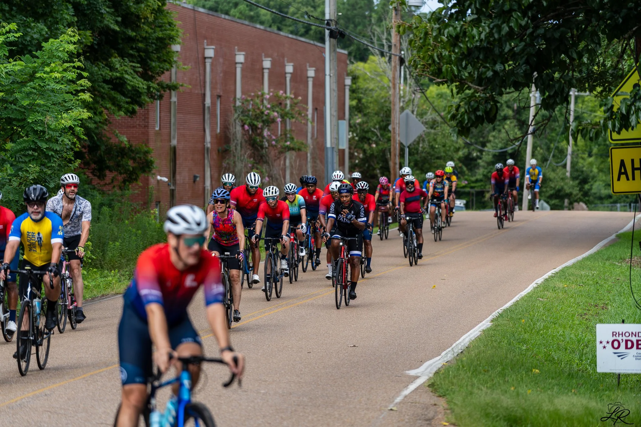 A large group of cyclists participating in a race or event on a suburban street surrounded by trees and buildings, with some wearing colorful helmets and jerseys.