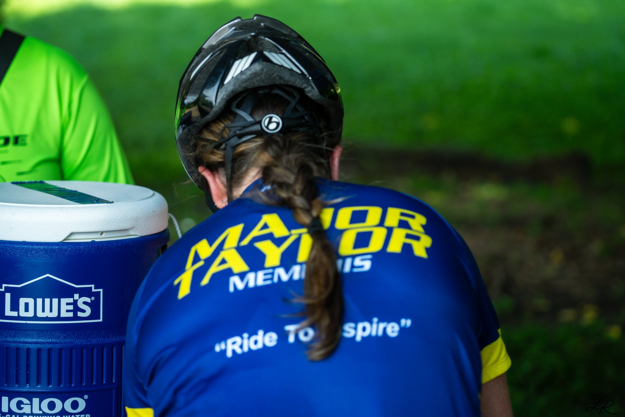 Back of a female cyclist wearing a blue jersey with yellow accents, a black helmet, and braided hair. She is sitting next to a white and blue cooler with the Lowe's logo. The background is green with grass or trees.
