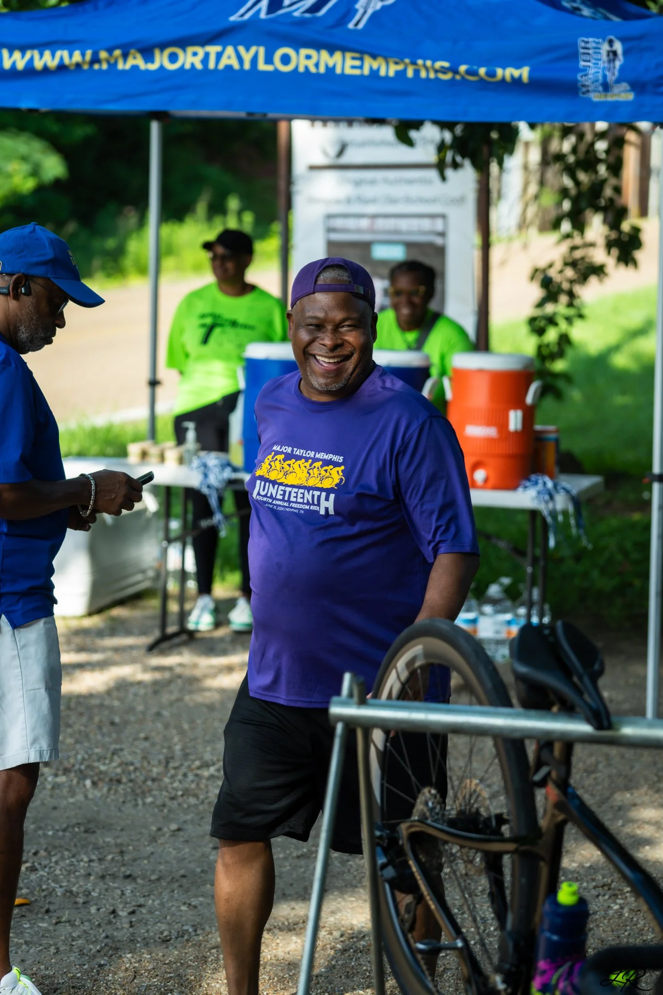 A smiling man in a purple shirt and purple cap standing outdoors near a wheelchair. There are people wearing bright green shirts behind a table, with a blue canopy overhead that has a website URL. The setting appears to be a community event or outdoo