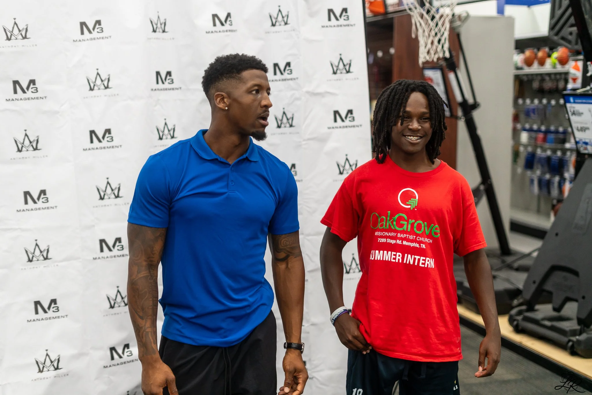 Two young men standing indoors with a white backdrop featuring the logo 'M3 Management'. One man wears a blue polo shirt with tattoos on his arms, the other wears a red t-shirt with 'OakGrove Missionary Baptist Church' printed on it, smiling. The bac