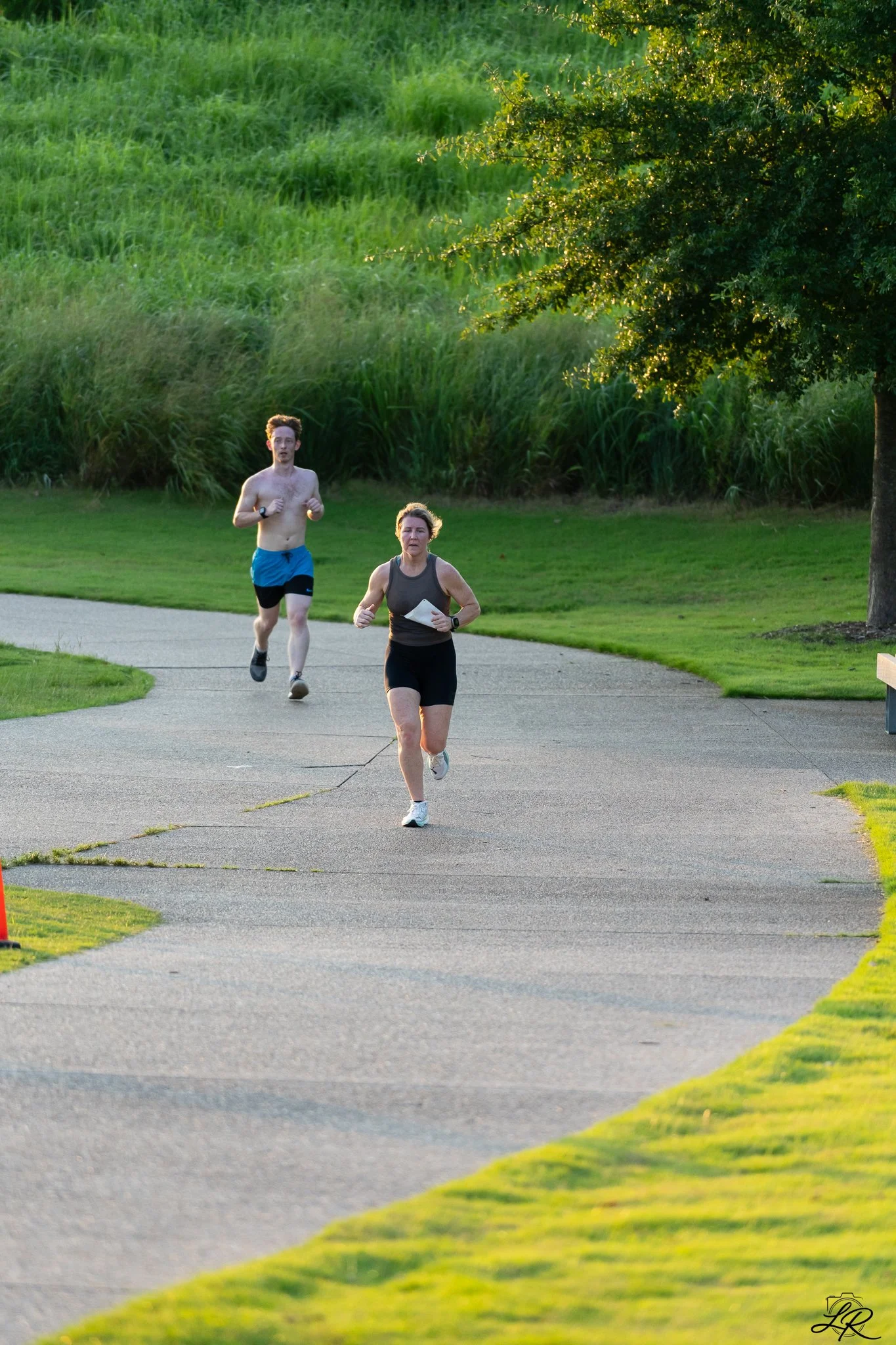 Two people running on a paved trail in a park, with green grass and a large tree on the right side of the image. The sky appears clear, suggesting a pleasant day.