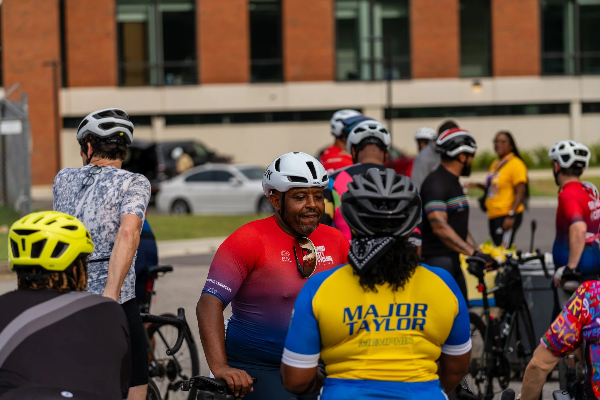 A group of cyclists gathered together, wearing helmets and colorful cycling jerseys, standing outdoors in an urban area with buildings and parked cars in the background.