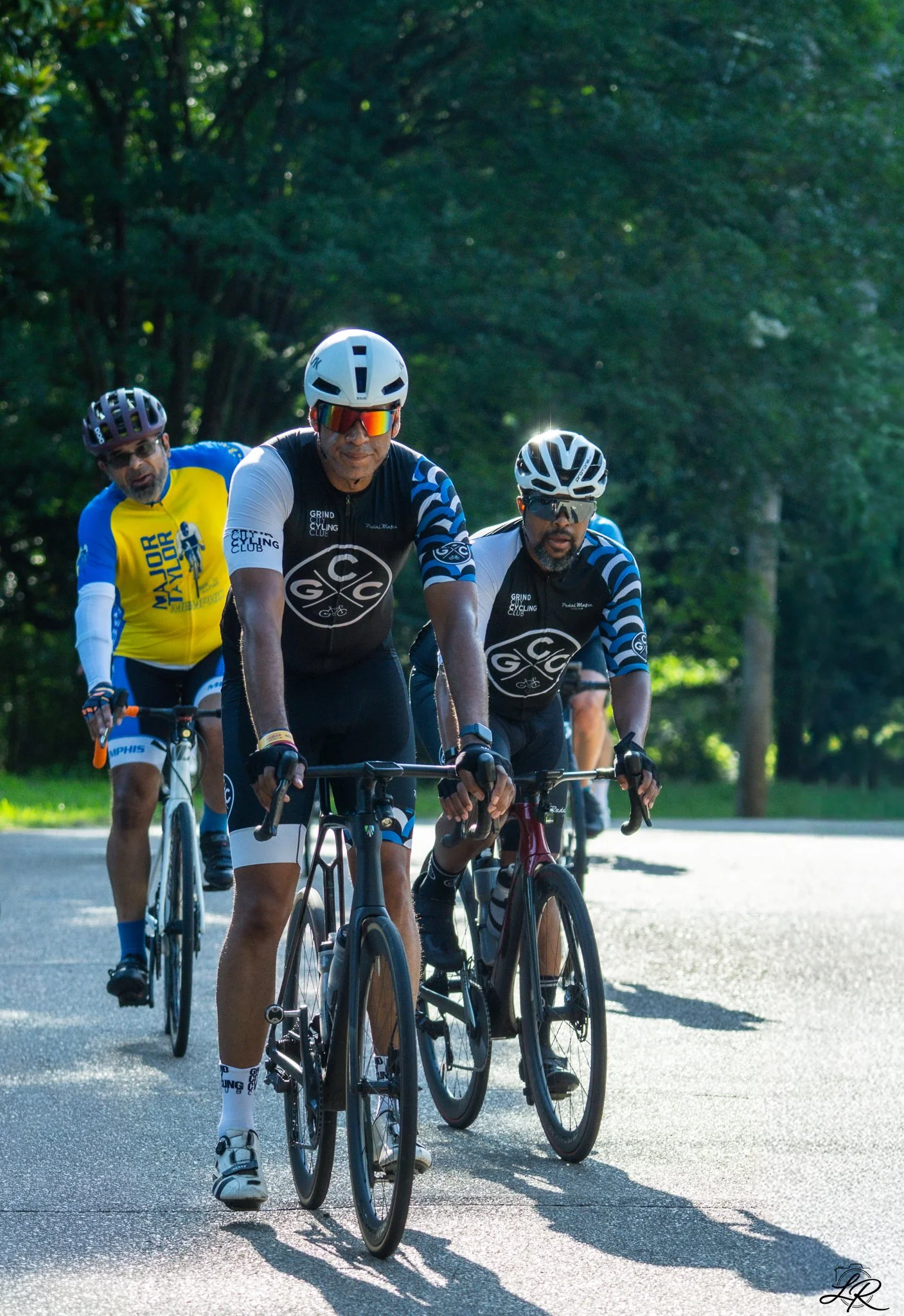 Three cyclists riding on a road surrounded by trees, wearing helmets and cycling gear, with sunlight creating shadows on the asphalt.