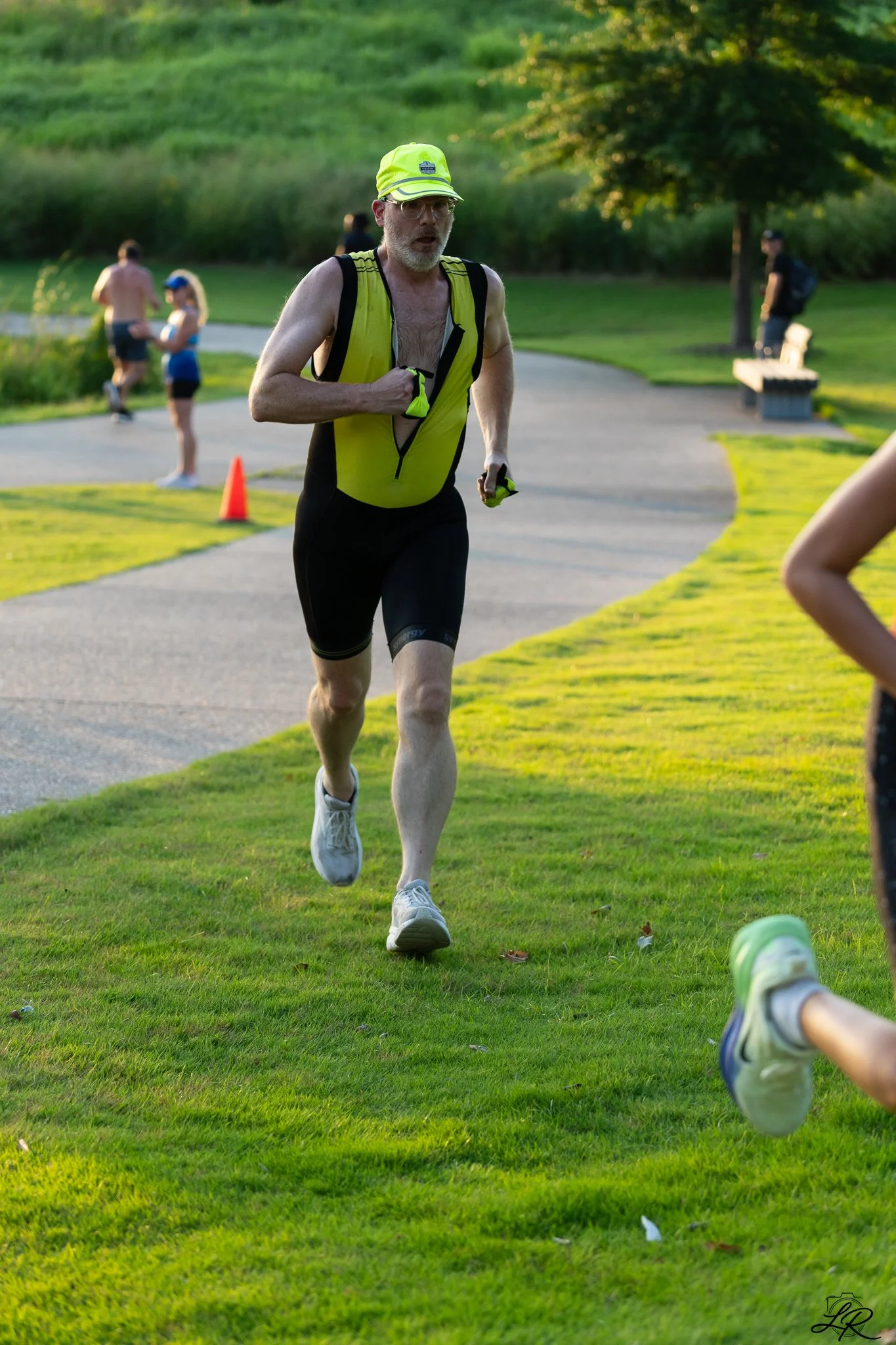 A man running in a park during sunset, wearing a yellow and black athletic outfit, including a hat and gloves, with other park visitors in the background.