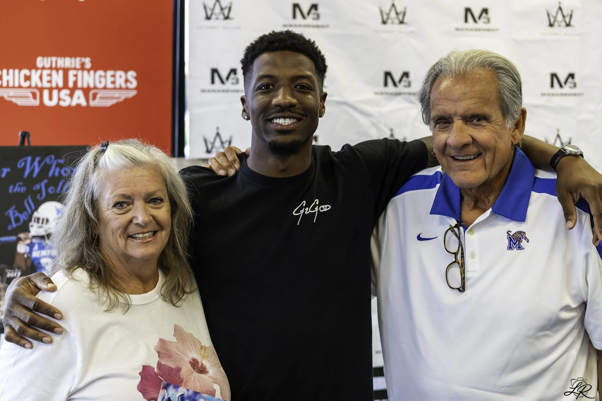 Three people, two women and a man, smiling and posing for a photo together with their arms around each other at an event. There is a red sign with white text and a white background with black logos behind them.