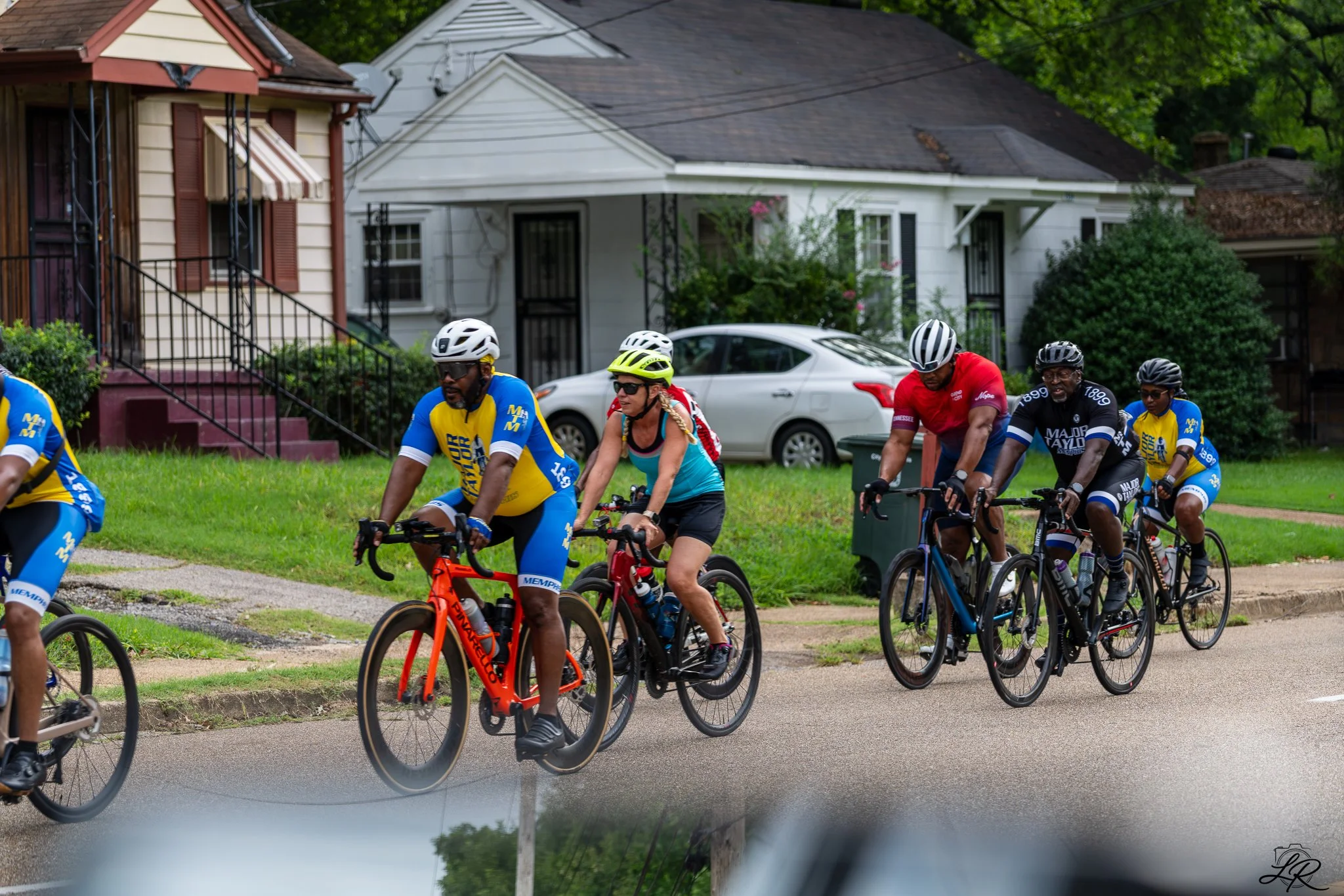 A group of people riding bikes in a neighborhood with houses and greenery in the background.