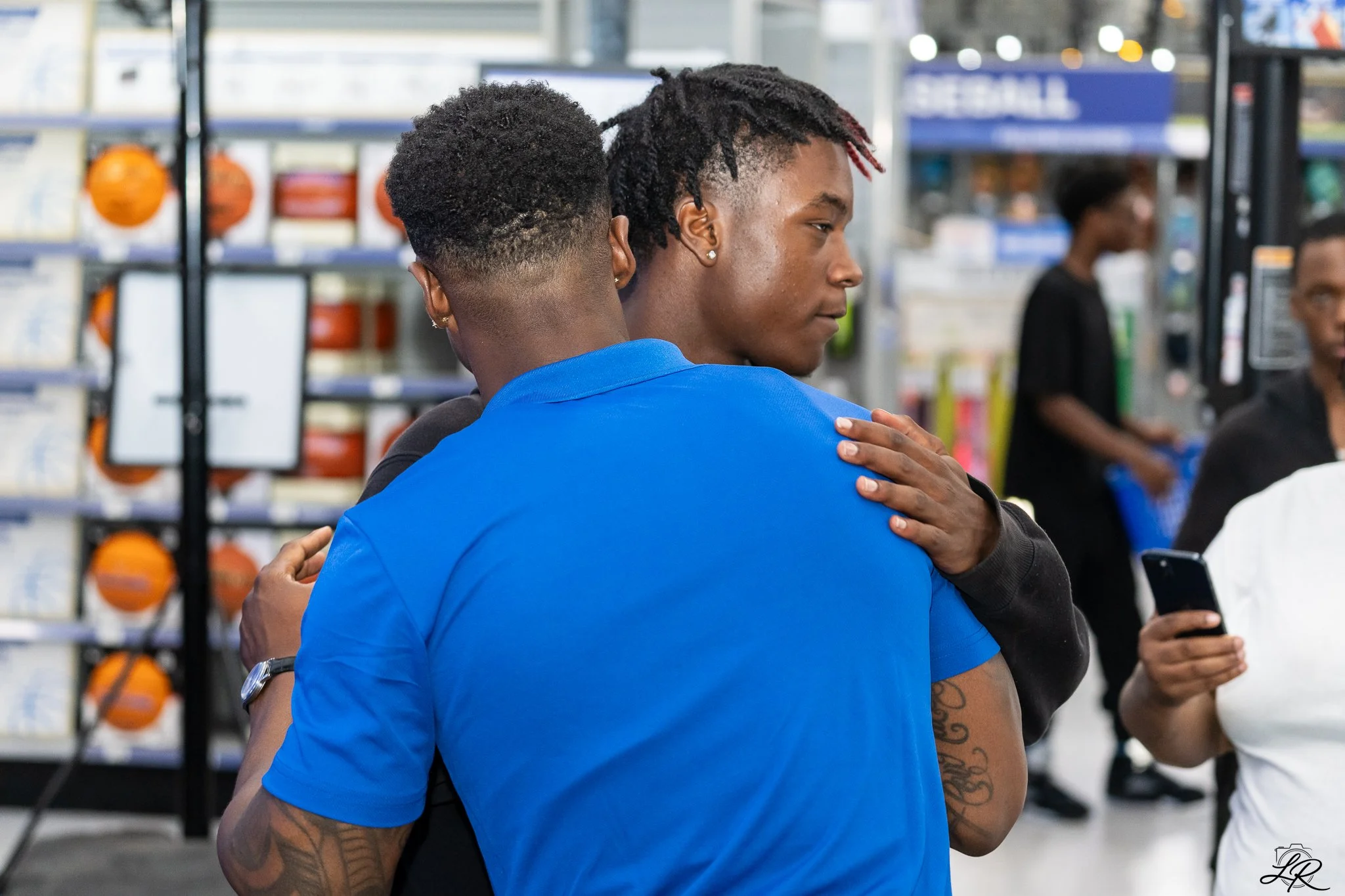 Two men with dark curly hairstyles embrace inside a store with shelves of basketballs in the background.