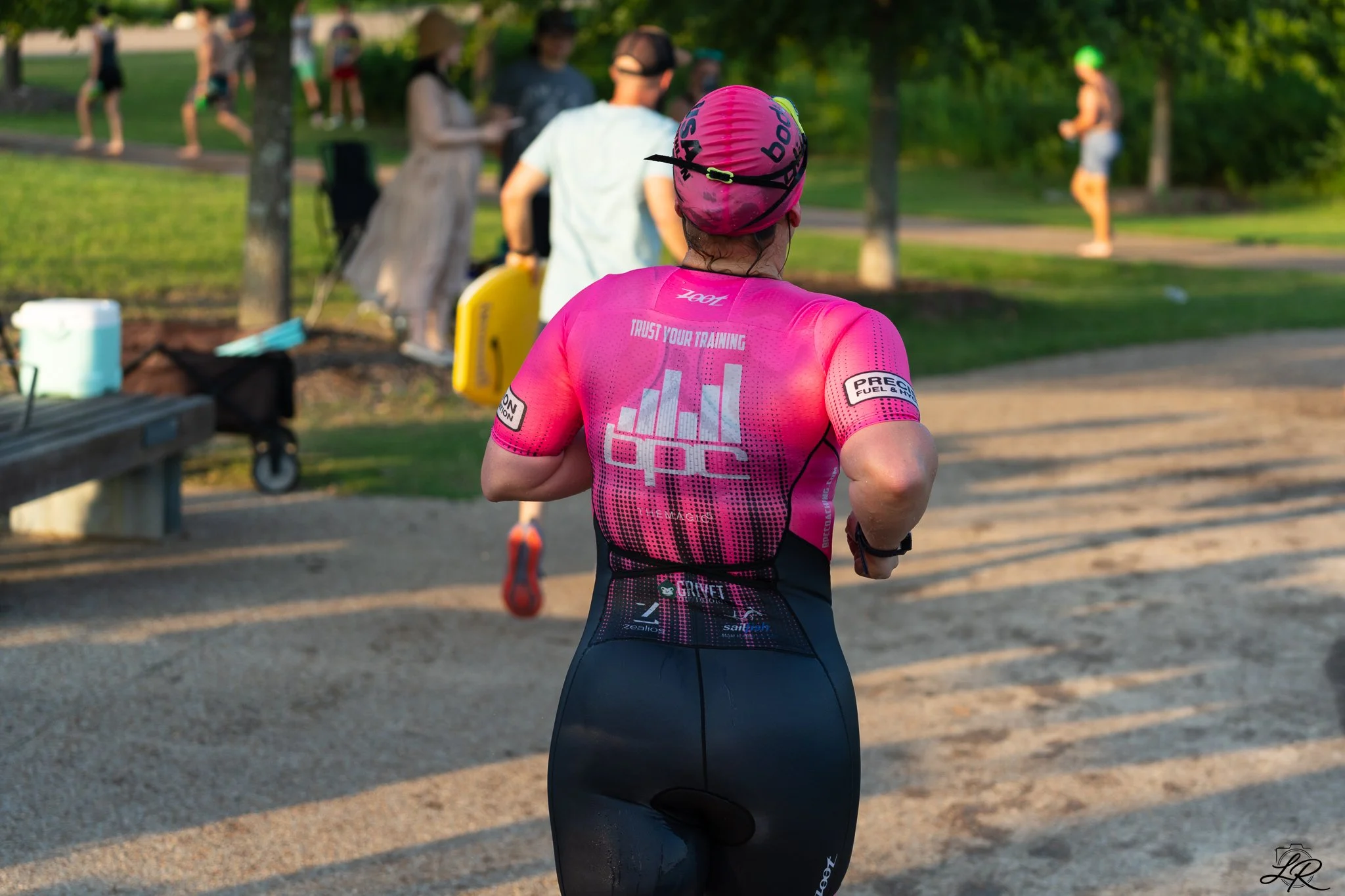 A woman wearing a pink athletic outfit and a pink cap running outdoors on a sunny day, with people and trees in the background.