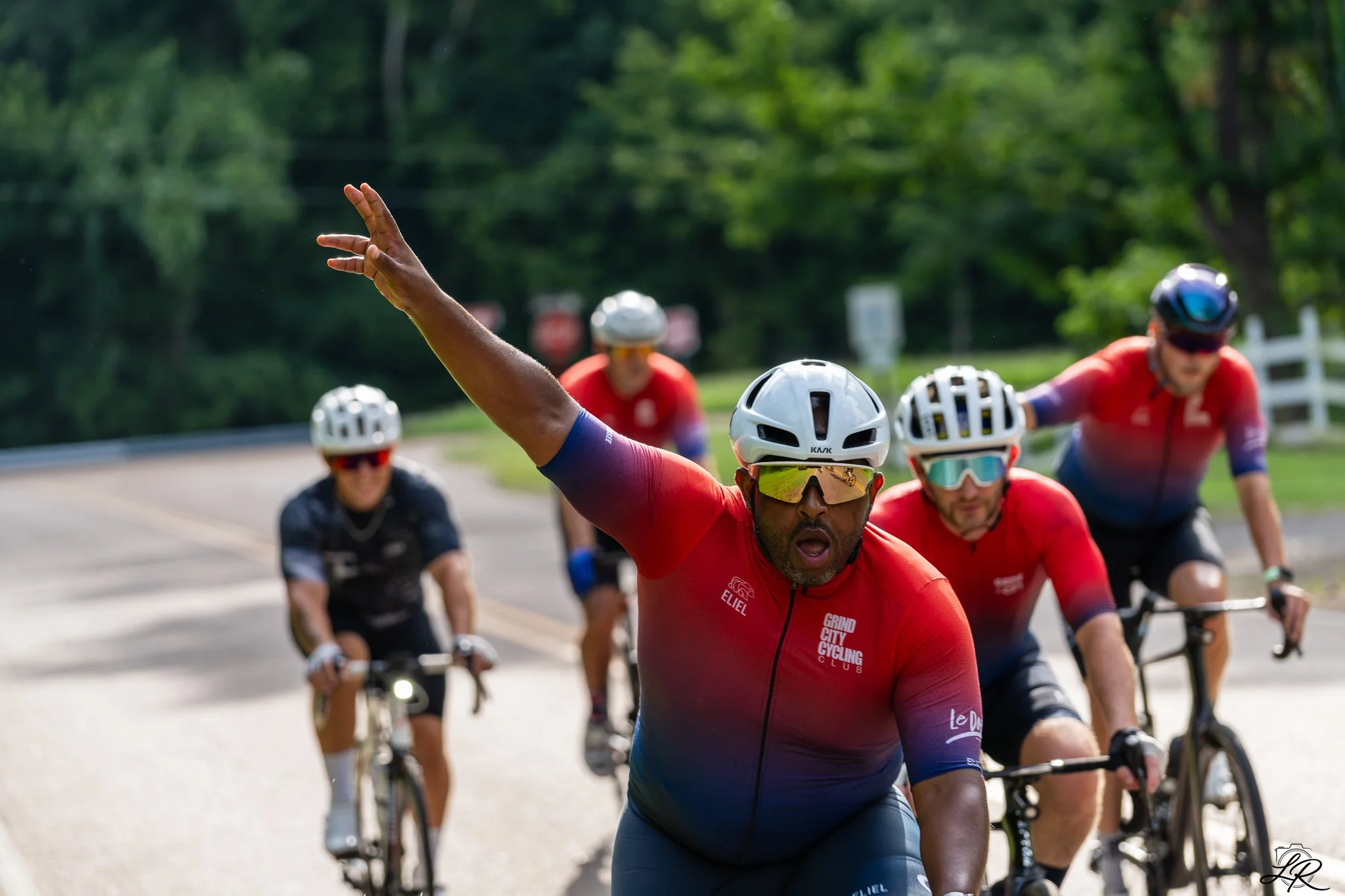 A group of cyclists wearing helmets and cycling jerseys riding on a road through a green, wooded area, with one cyclist in the foreground raising their hand.