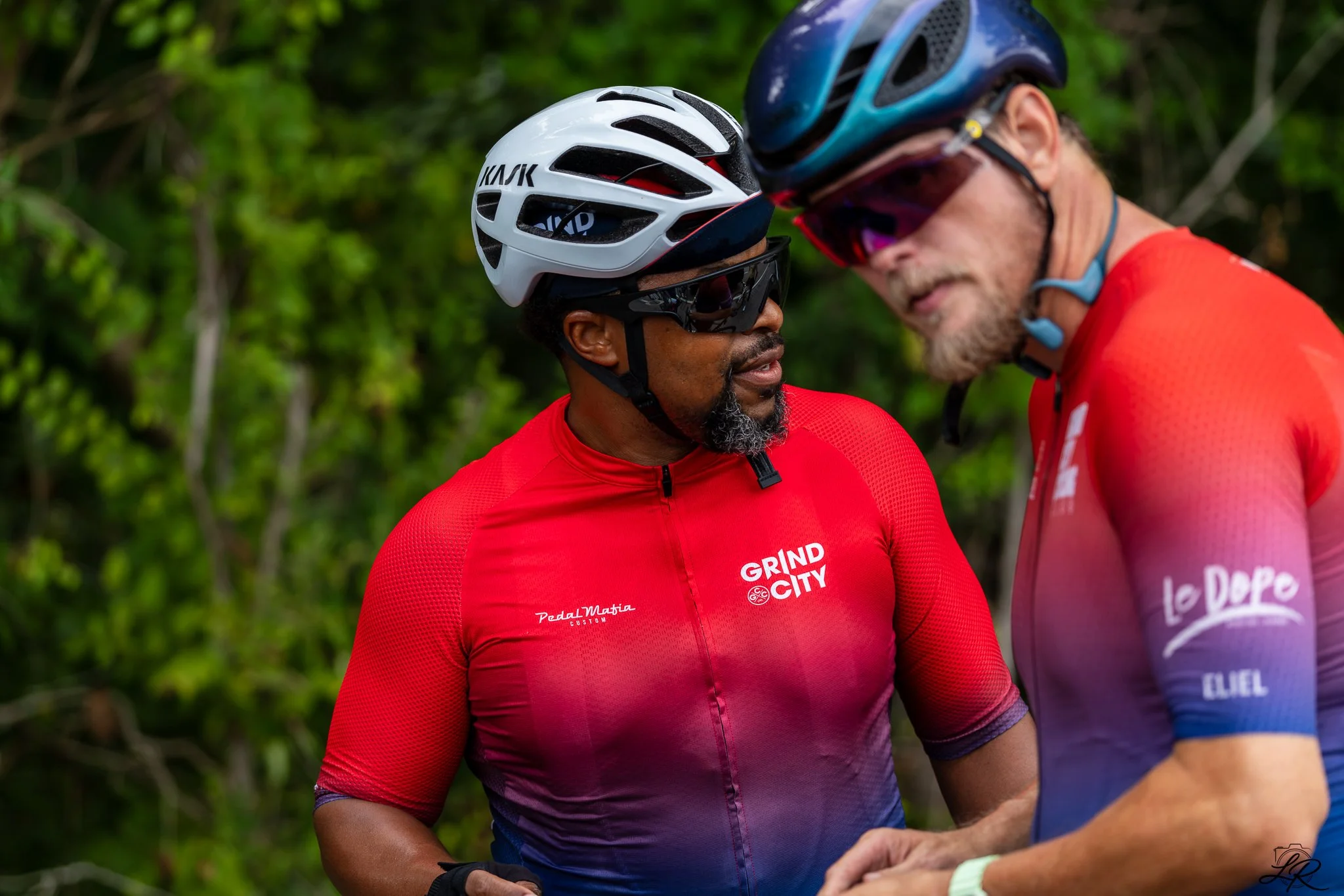Two cyclists wearing helmets and sunglasses have a conversation outdoors amid green foliage. One cyclist's red jersey has the words 'GRIND CITY' printed on it.