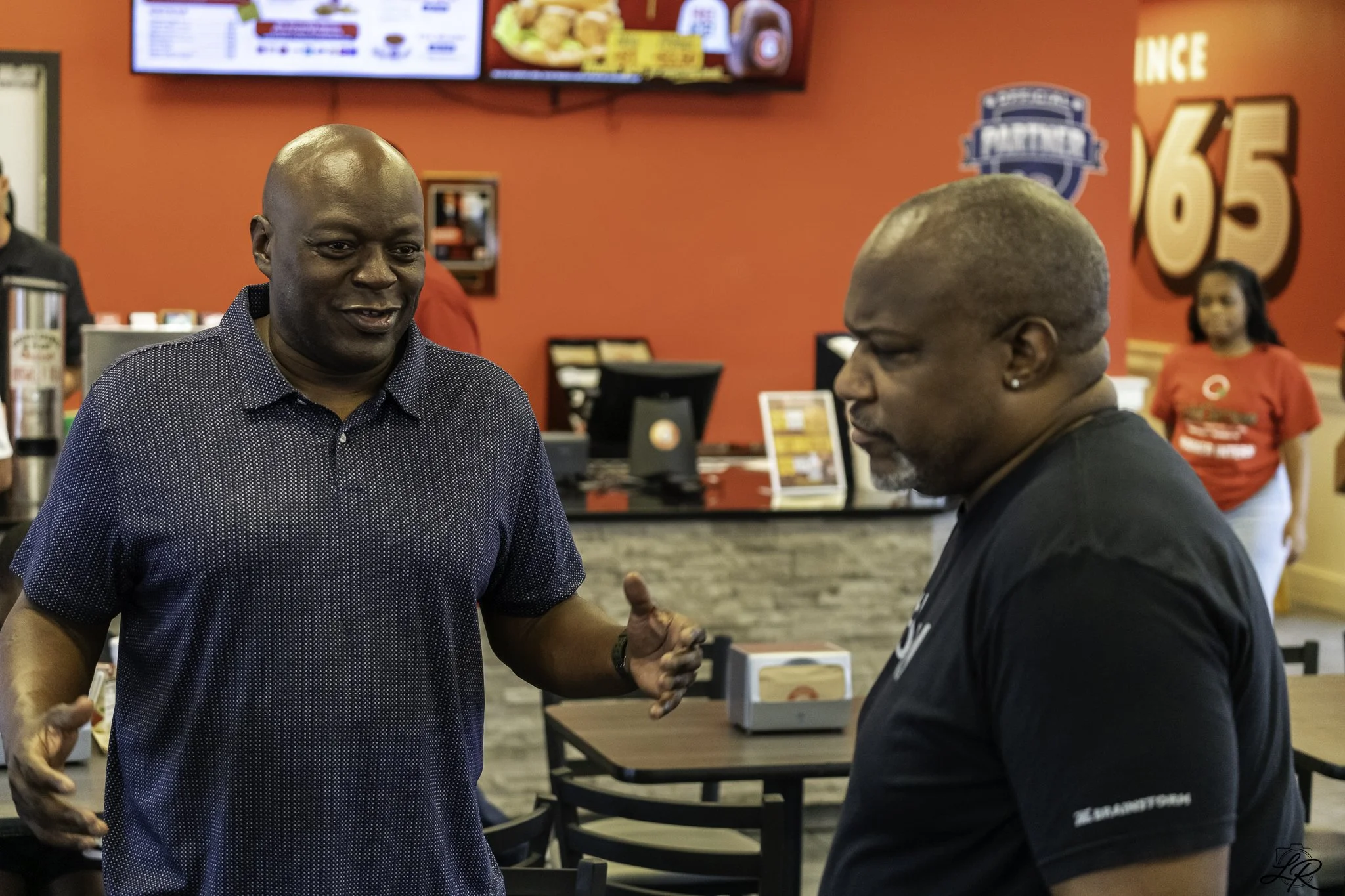Two men are shaking hands and conversing inside a fast-food restaurant with a red wall. One man is bald and wearing a dark, dotted shirt, while the other has short gray hair and wears a black T-shirt. In the background, there is a television, menu, a