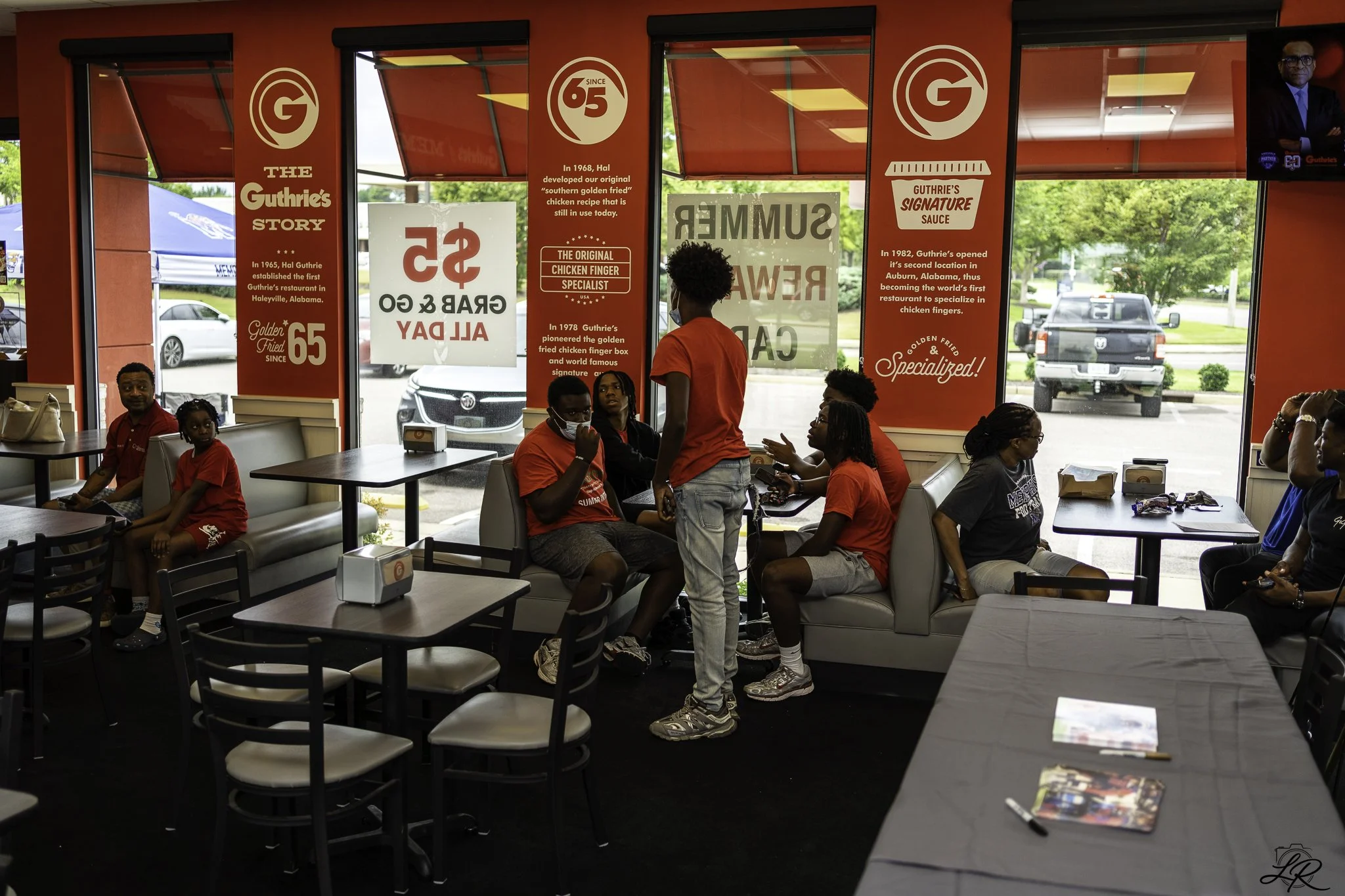 Inside a fast-food restaurant with several customers sitting at tables and a staff member standing and talking to a seated customer. There are large red windows with signage describing the restaurant's history and signature chicken recipes. Outside, 