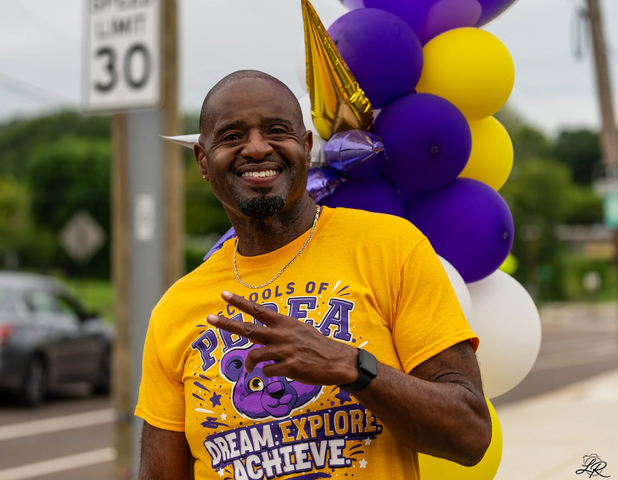 Smiling man with tattoos wearing a yellow T-shirt standing outdoors with purple and yellow balloons behind him.