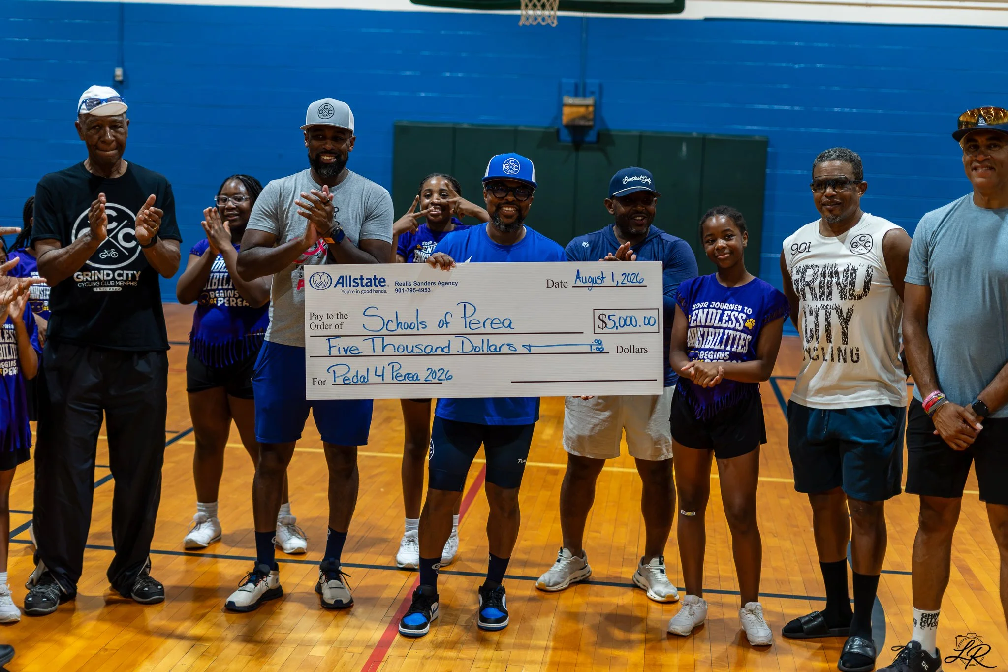 A group of diverse people, including children and adults, are standing on a basketball court inside a gymnasium, holding an oversized check for $5,000 made out to Schools of Perea for Pedal 4 Perea 2026. They are smiling, clapping, and posing for the