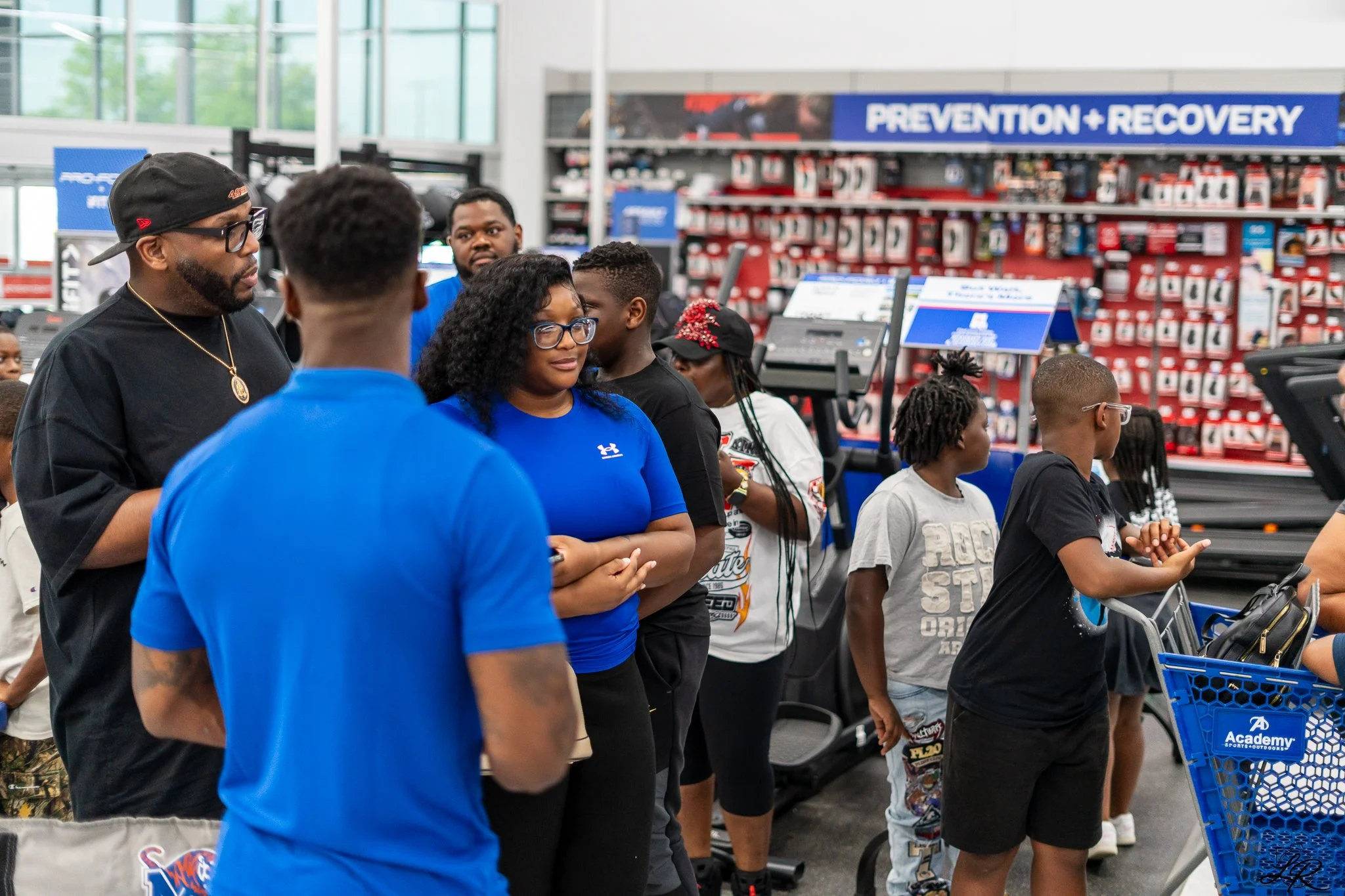 People standing in line inside a store with shelves of automotive supplies in the background, including a sign reading 'Prevention + Recovery'.