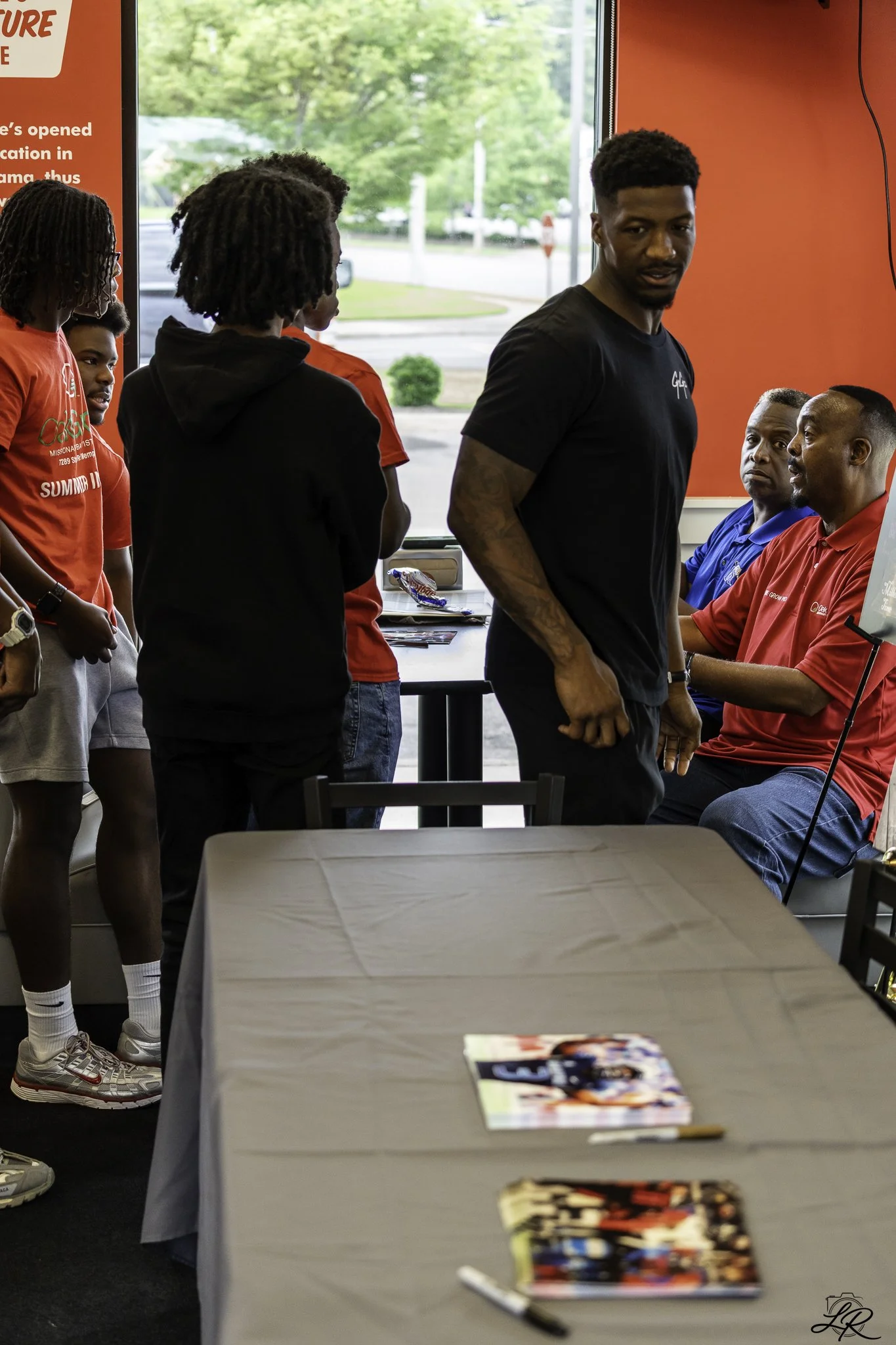 Inside a fast food restaurant with a group of African American teenagers standing in line. Two adults are seated at a table, one wearing a red shirt and the other in a blue shirt. The restaurant has red walls and a large window showing trees outside.