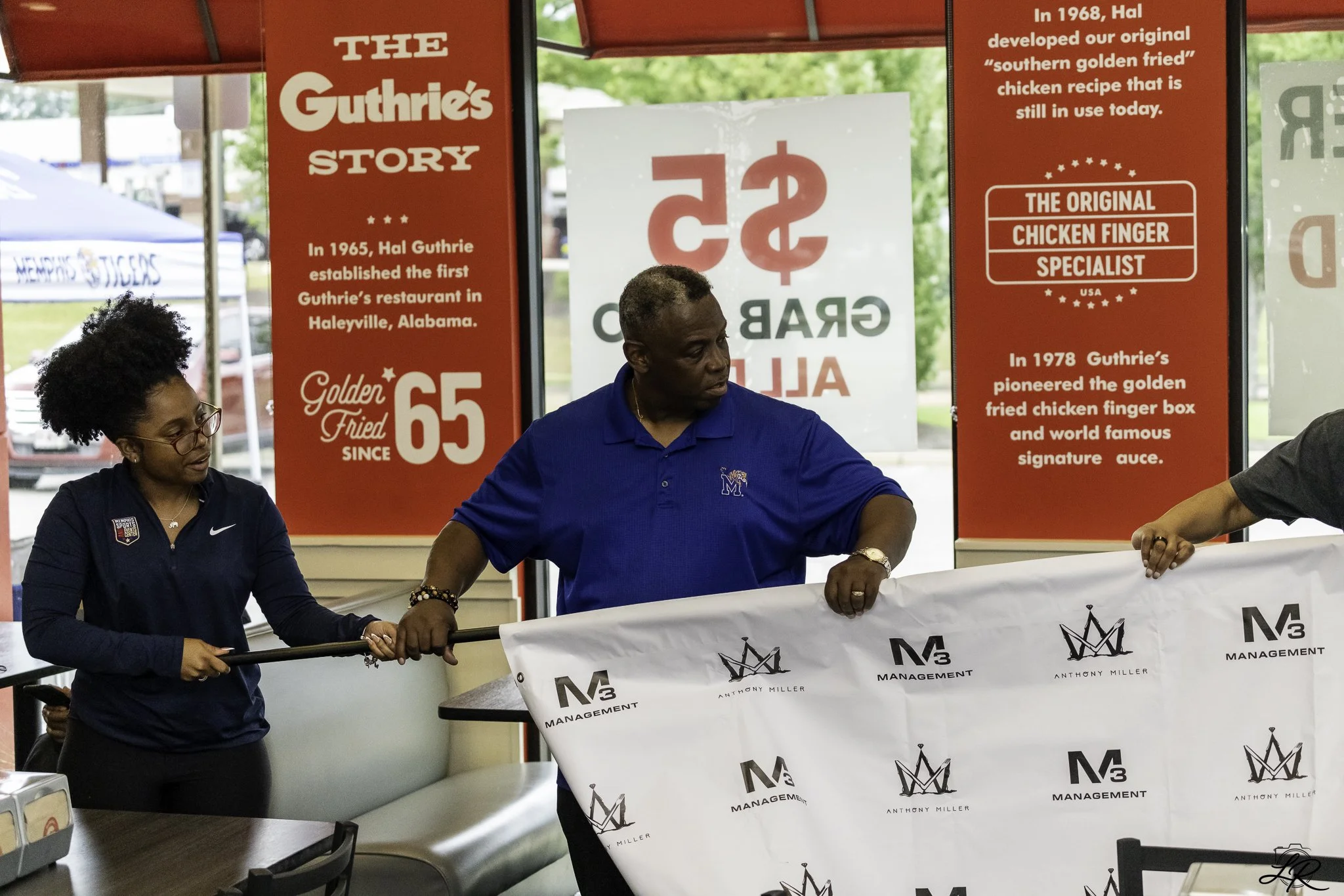Three people at a table holding a banner with the logos of M3 Management and Anthony Miller. Red and white informational banners are in the background, describing the history of Hal Guthrie's restaurant and famous chicken finger recipe.