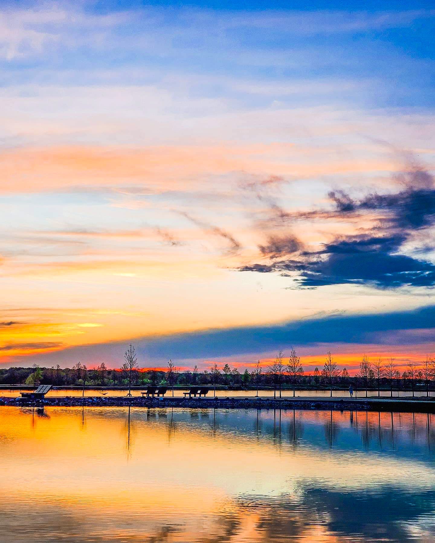 Sunset over a calm lake with a pier, trees, and benches reflected in the water, with colorful sky and clouds.
