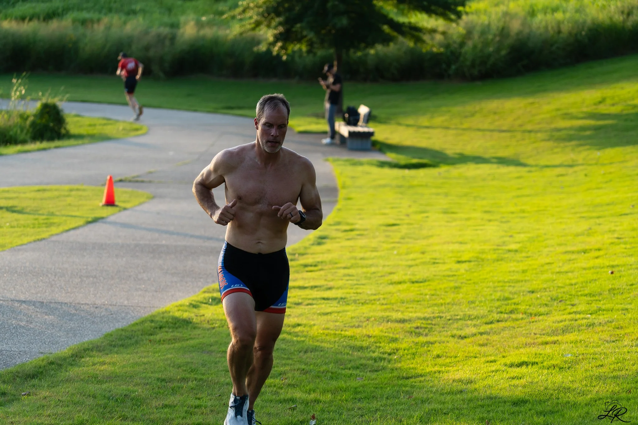 A shirtless man running on a paved path in a park surrounded by green grass and trees, with a person on a skateboard and another person standing near a bench in the background.