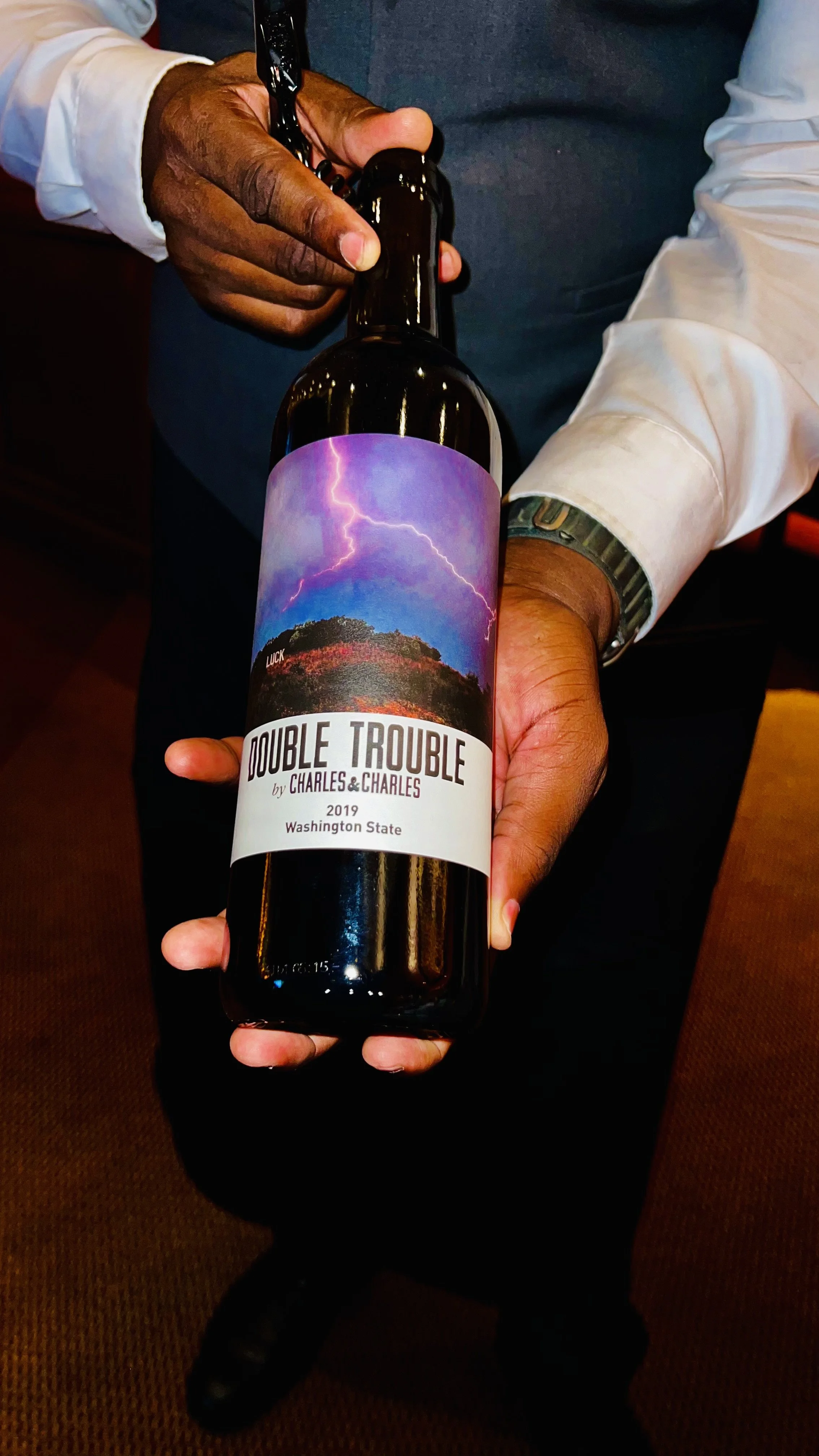 A person with dark skin and a white shirt holding a wine bottle labeled 'Double Trouble' with a scenic label featuring lightning and a mountain, against a background of brown carpet.