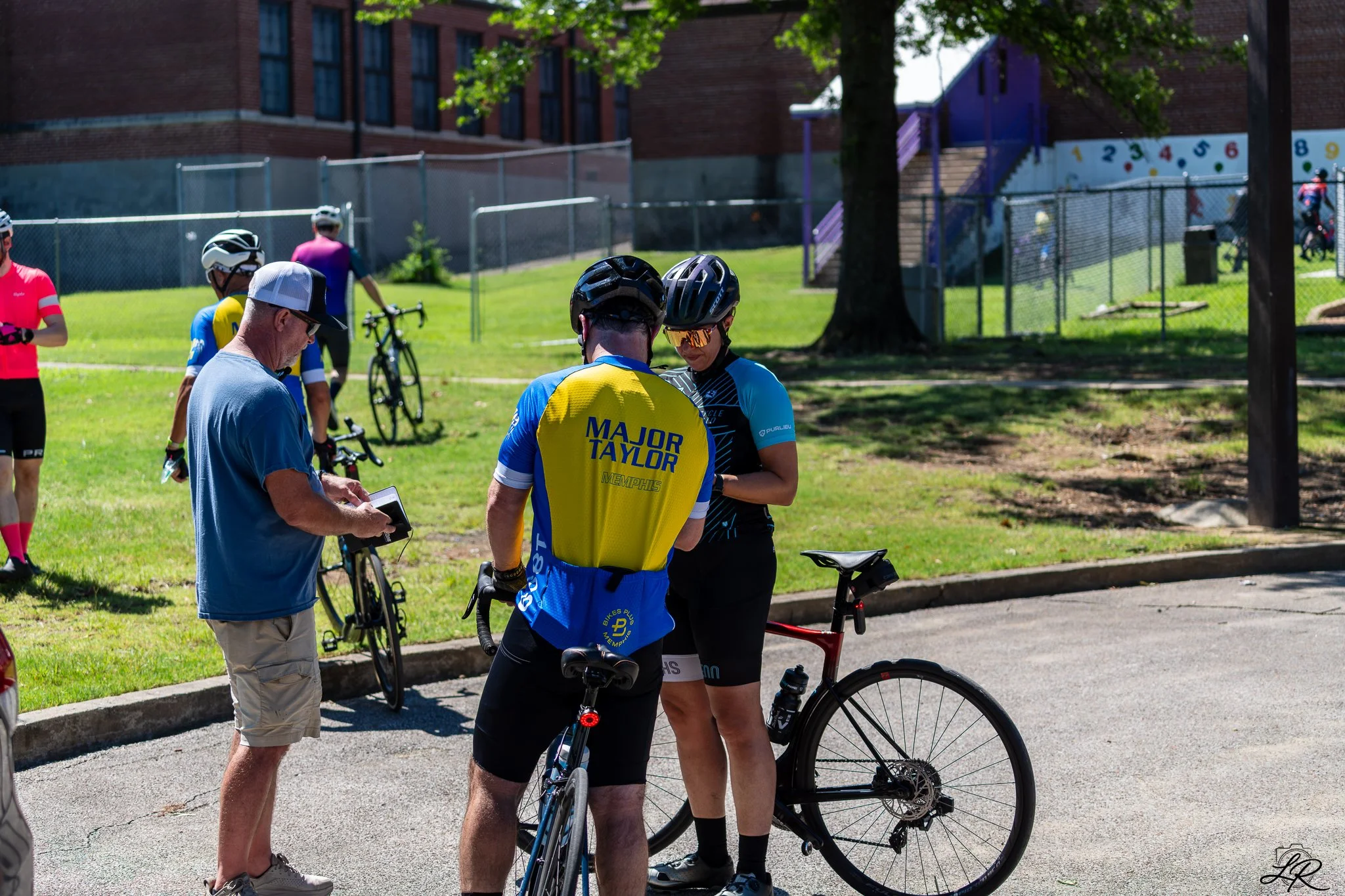 Group of cyclists and a man in a blue shirt standing outdoors on a sunny day, with some people wearing helmets, near a brick building and a playground area.
