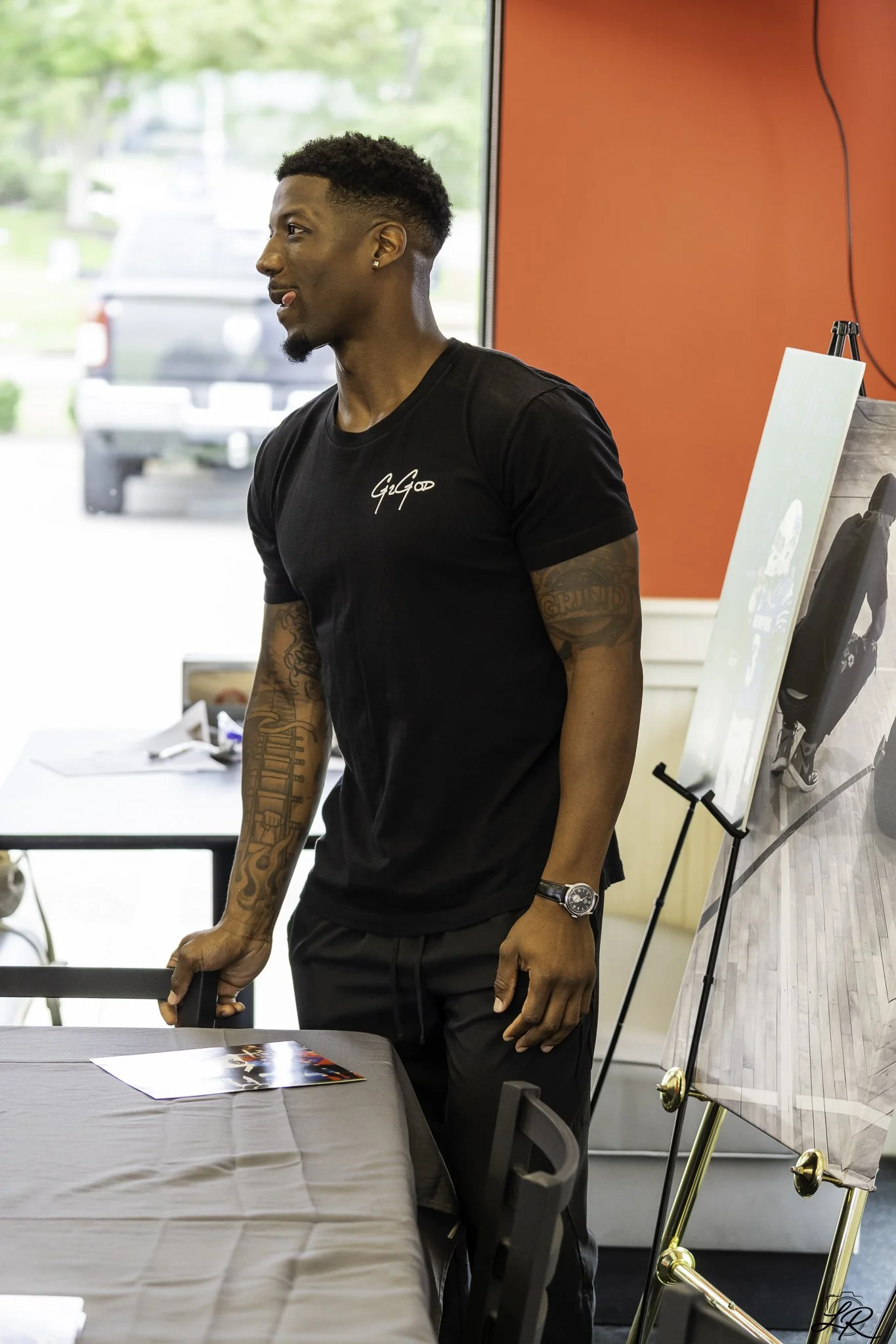 A young man with tattoos wearing a black T-shirt with a signature logo, standing in a room with bright natural light, holding a black object while looking to the side. There is a table with papers and photos, and an easel with a canvas behind him.