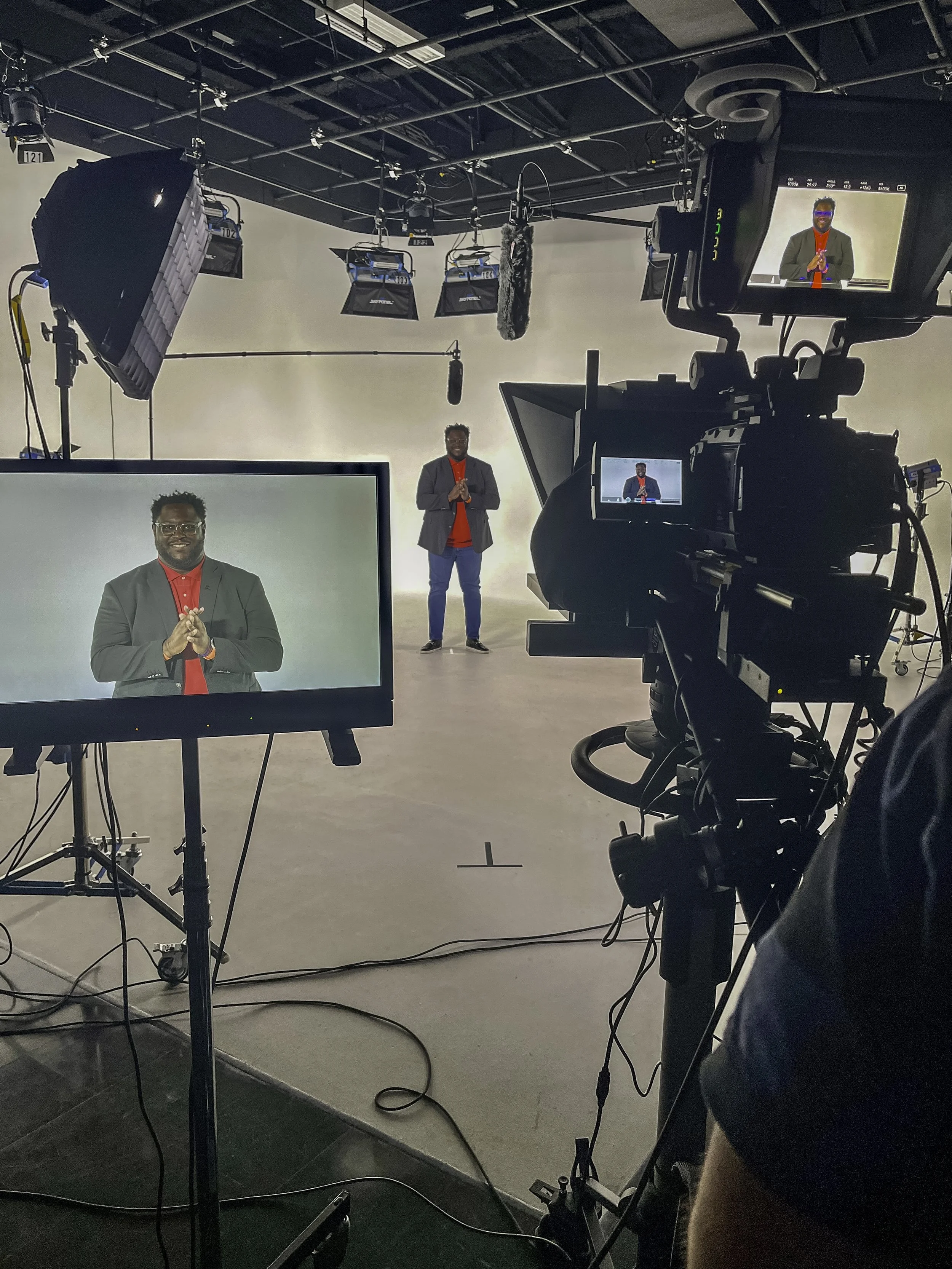 A video recording studio with a person standing in front of a white backdrop, studio lights, a camera, and monitors showing the person. The person is wearing a gray blazer over a red shirt, and they are smiling with hands clasped.