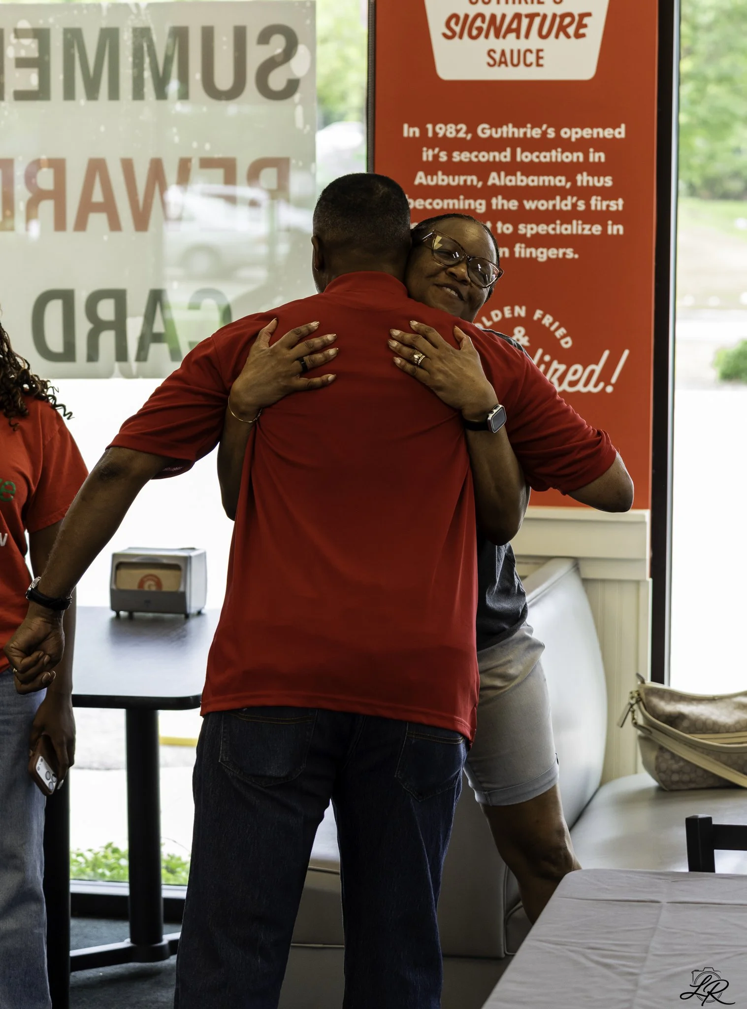Two people hugging inside a restaurant, with a red and white sign showing the history of Guthrie's sauce in the background.