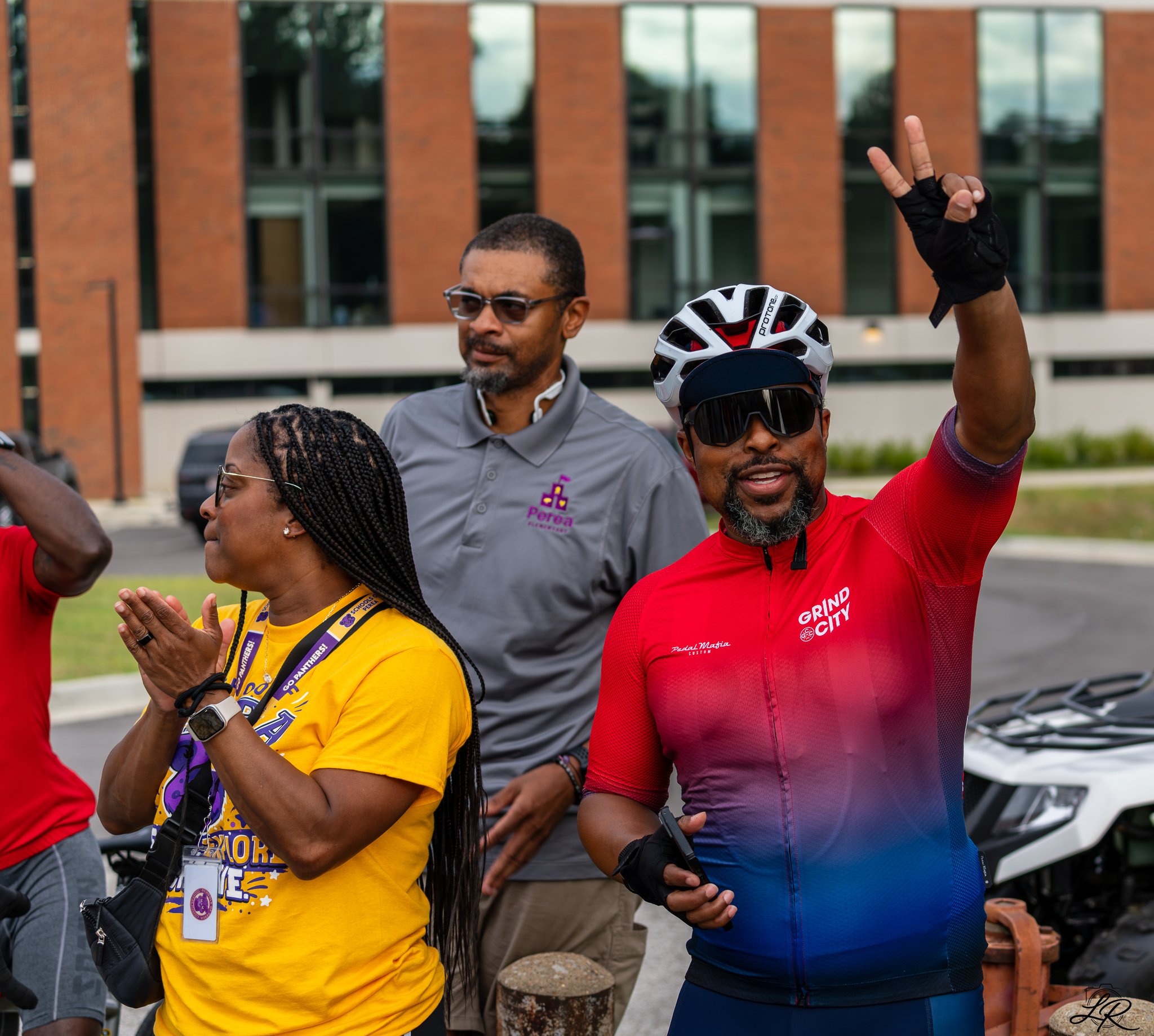 A group of diverse people gathered outdoors, some wearing cycling helmets, with one person raising two fingers in a peace sign. The background shows a modern brick building with large windows.