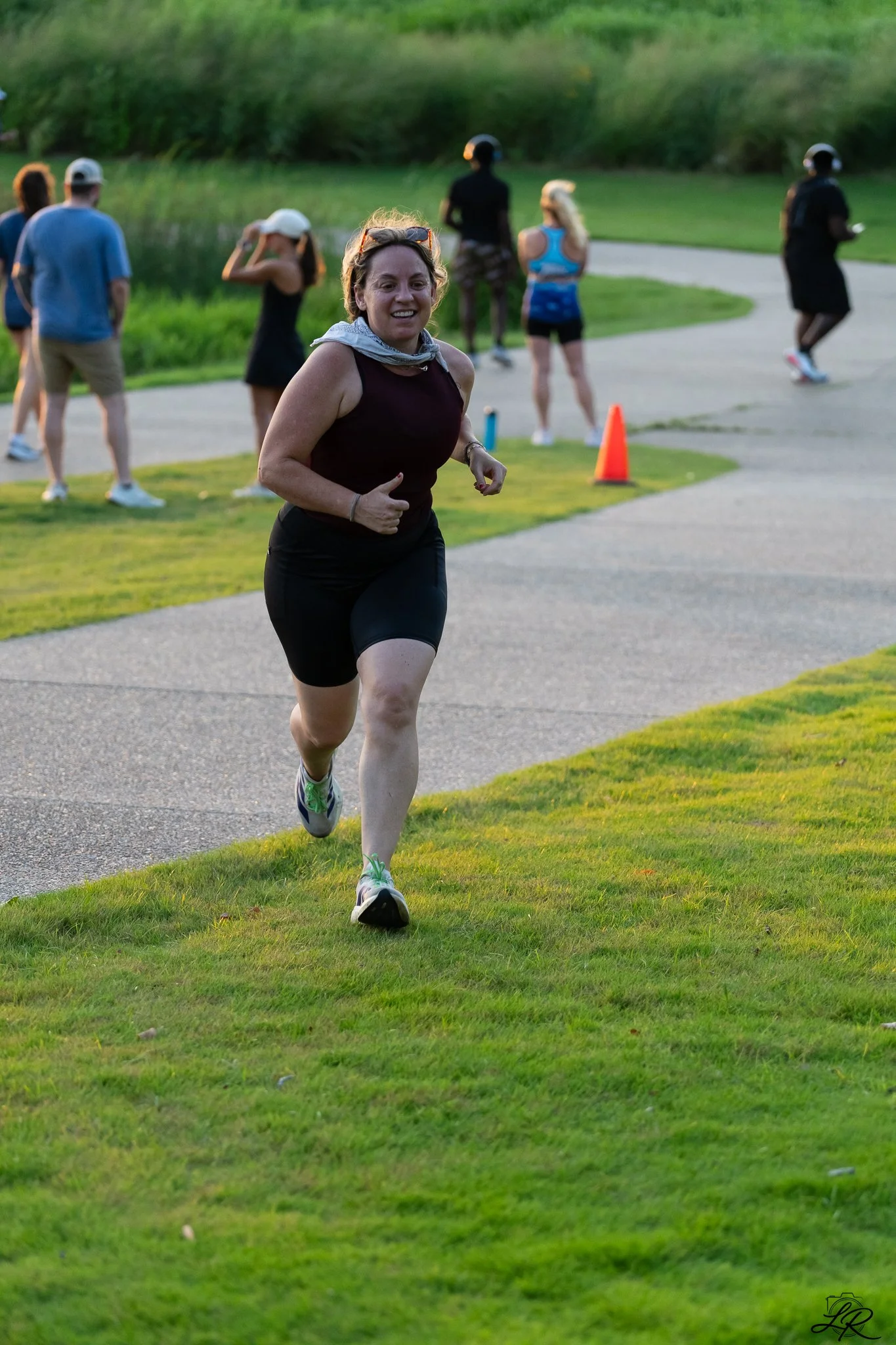 A woman running outdoors on a grassy area with trees and a pond in the background. She is wearing a black running outfit, a gray scarf, and running shoes. Other people are standing and walking nearby, some wearing helmets and athletic clothing.
