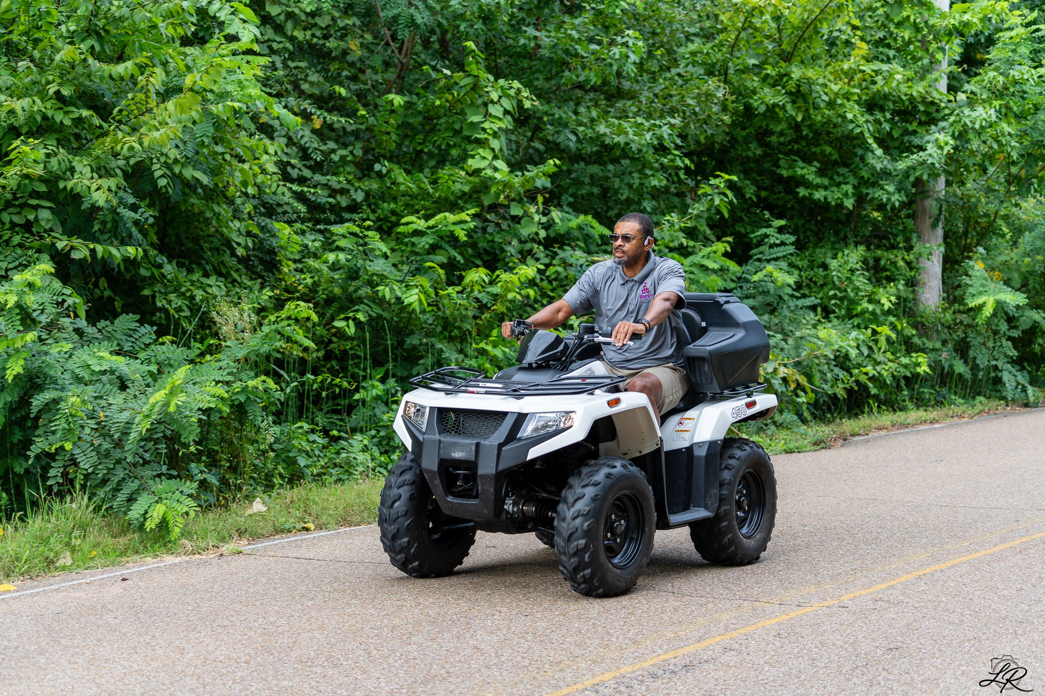 A man riding an all-terrain vehicle (ATV) along a paved road next to a green, leafy forest.