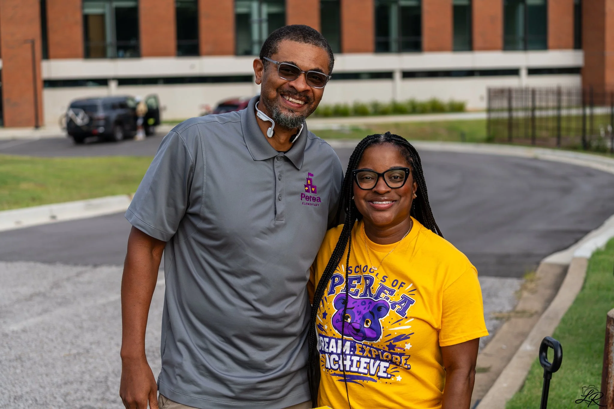 Smiling man and woman standing outdoors in front of a school building, both wearing glasses and casual clothing, with the man in a gray shirt and the woman in a yellow t-shirt promoting a school event.