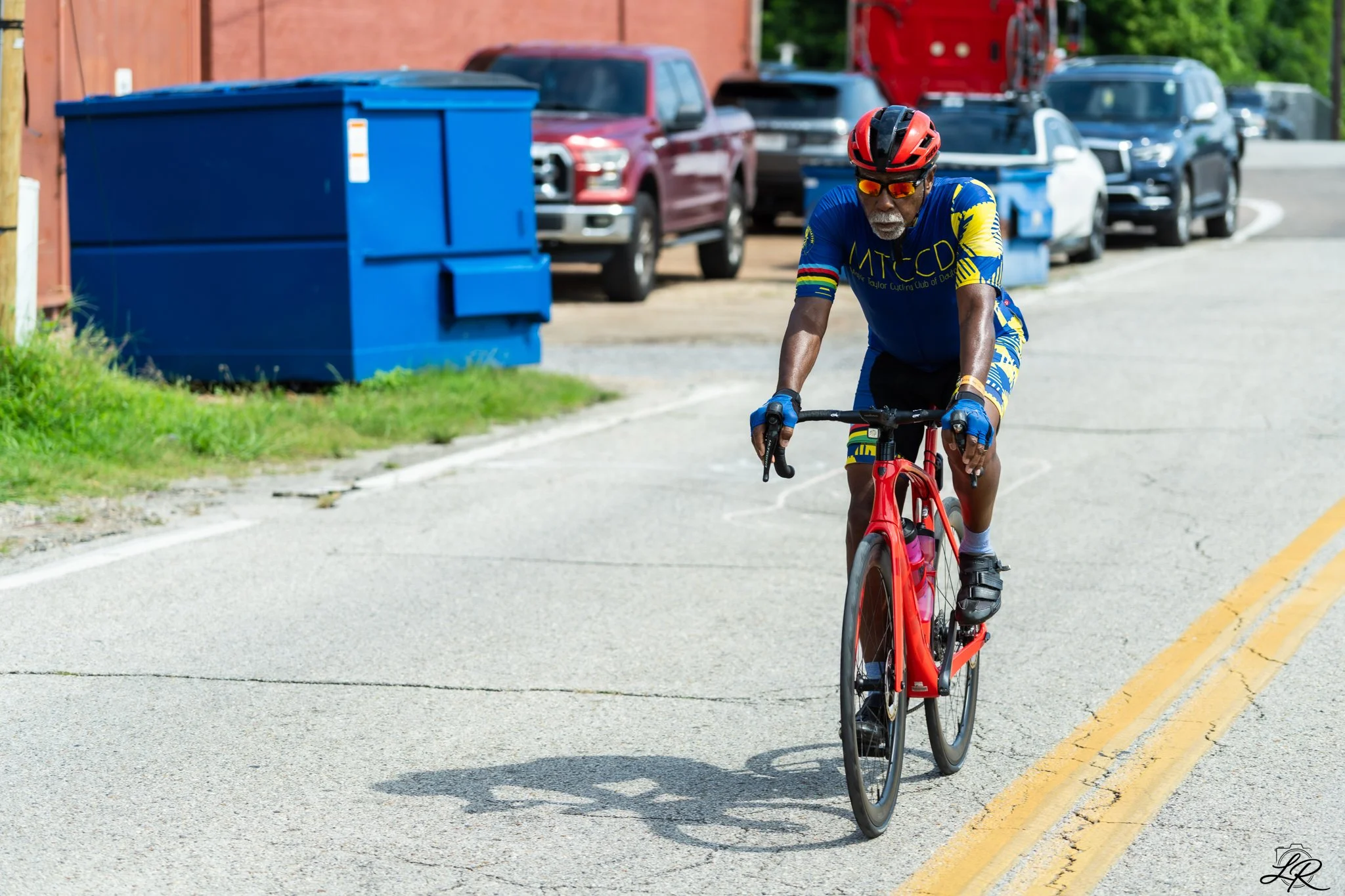 An elderly man with a gray beard, wearing sunglasses and a red helmet, riding a red road bicycle on a paved street. He's wearing a blue cycling jersey with yellow and red accents and black cycling shorts. Cars are parked along the side of the street 