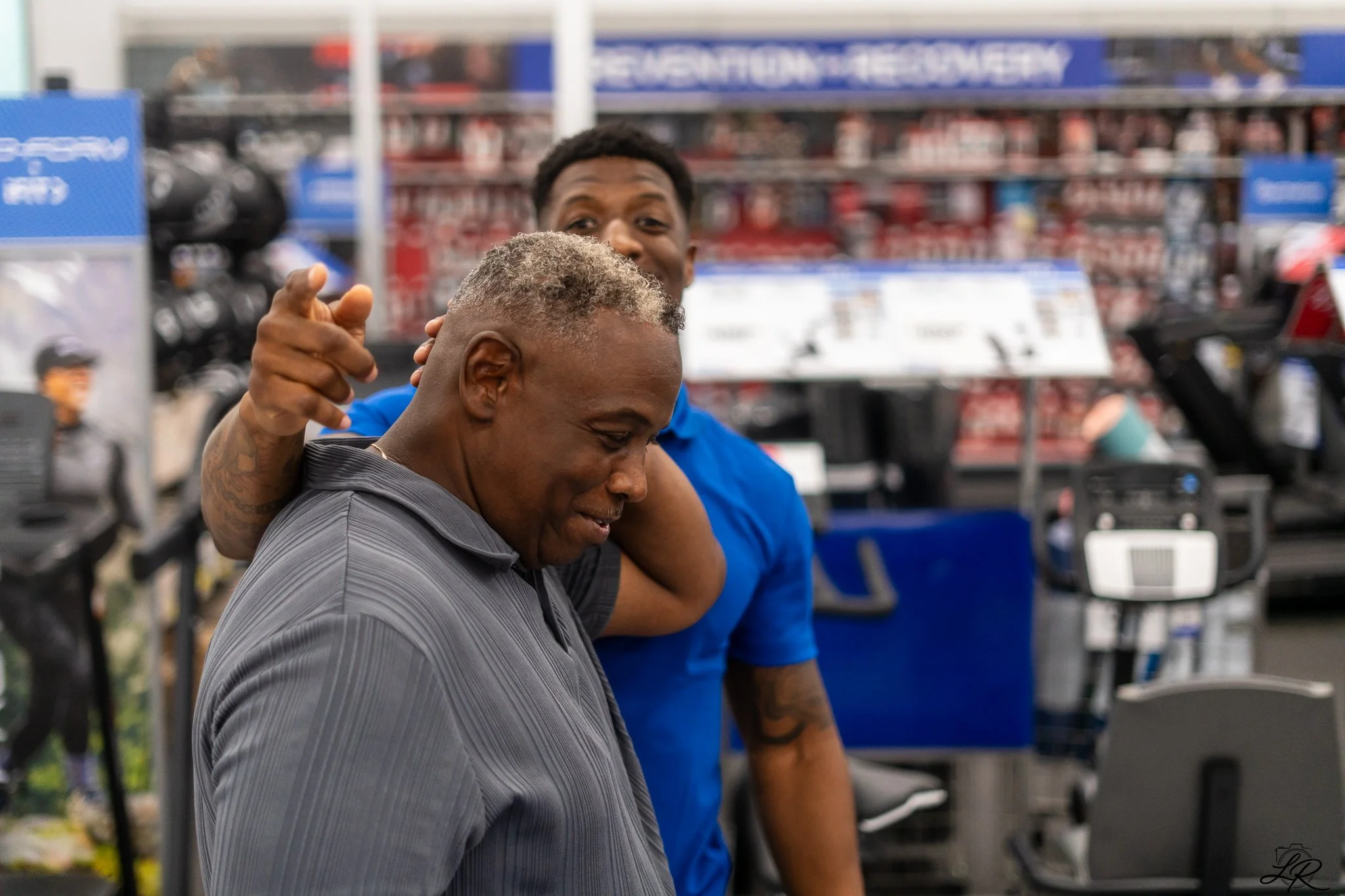 A man with short gray hair wearing a gray shirt stands in front of a younger man with short black hair in a blue shirt, inside an automotive store, with shelves of products and equipment in the background.